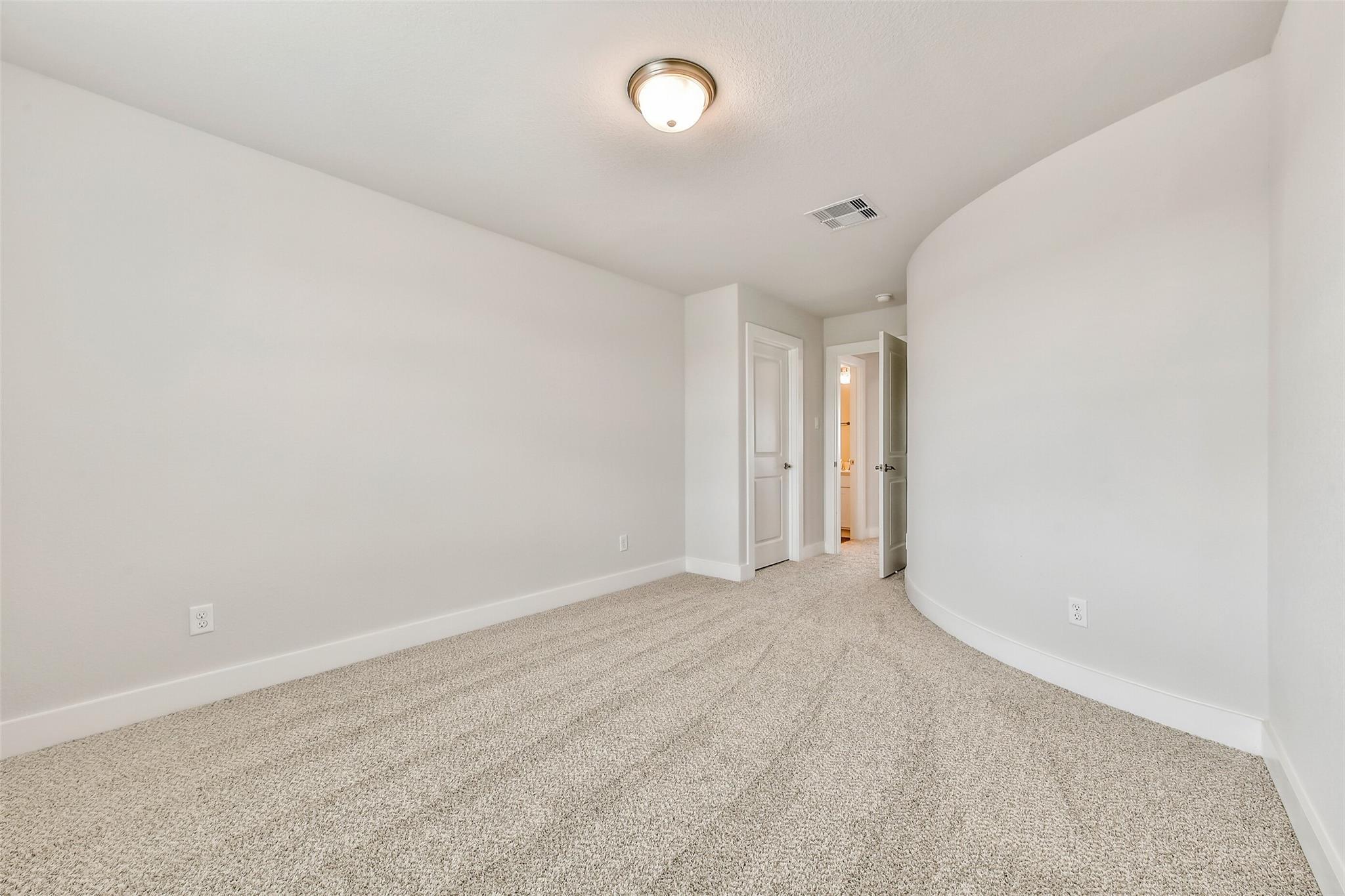 Spacious bedroom with curved white wall, beige carpet, and en-suite bathroom door in Davidson Homes The Victoria A, Lago Mar, Texas City