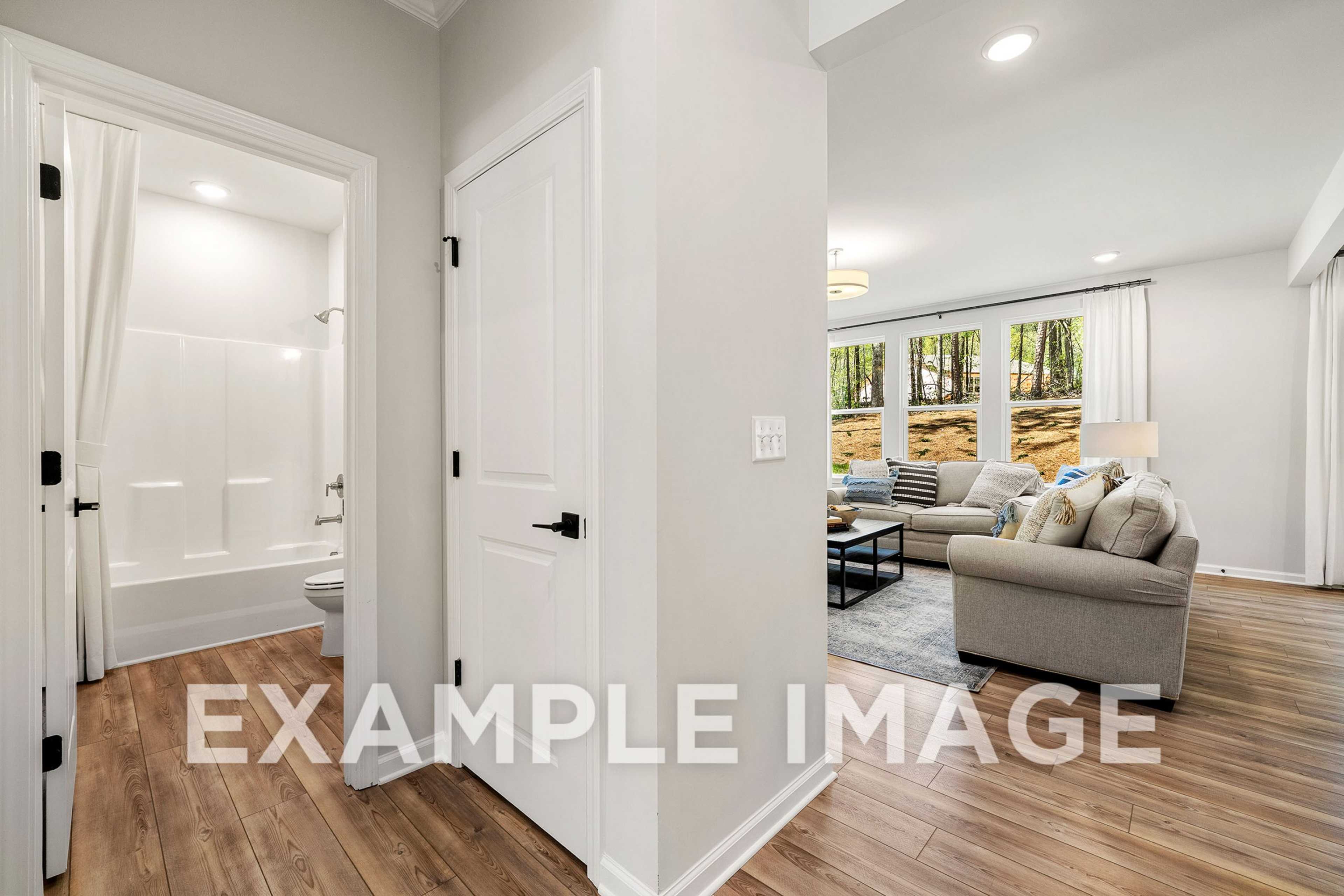 Interior of The Hickory A home design featuring white bathtub bathroom and open living room with beige sofa, wood floors, large windows