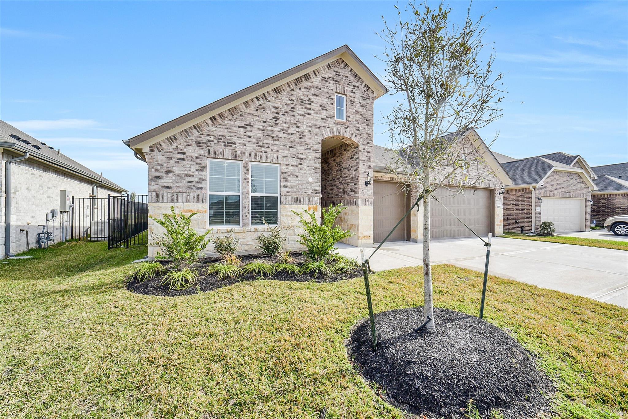 Brick single-story home with 3-car garage, young tree, shrubs, and manicured lawn in Sierra Vista, Rosharon, Texas