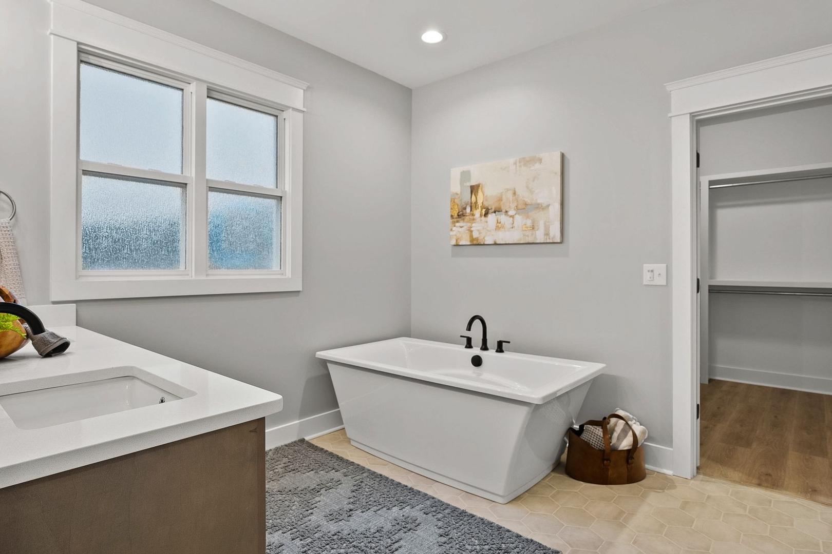 Spacious master bathroom in The Oxford with freestanding white tub, modern dark vanity, frosted window, and neutral gray tones