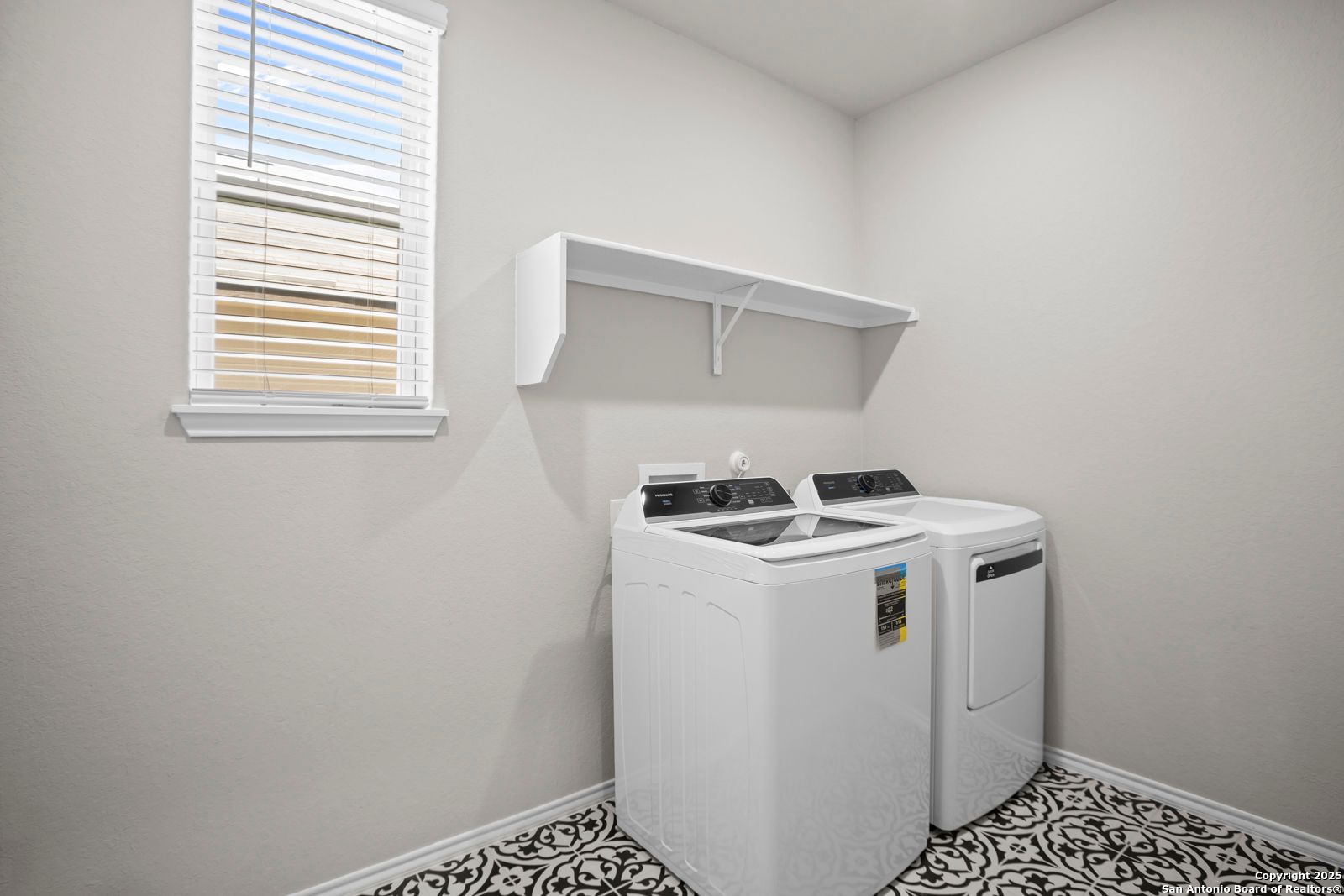 Modern laundry room with white washer-dryer set, floating shelf, blinds-covered window, and tiled floor in Davidson Homes Sequoia A, Converse, Texas