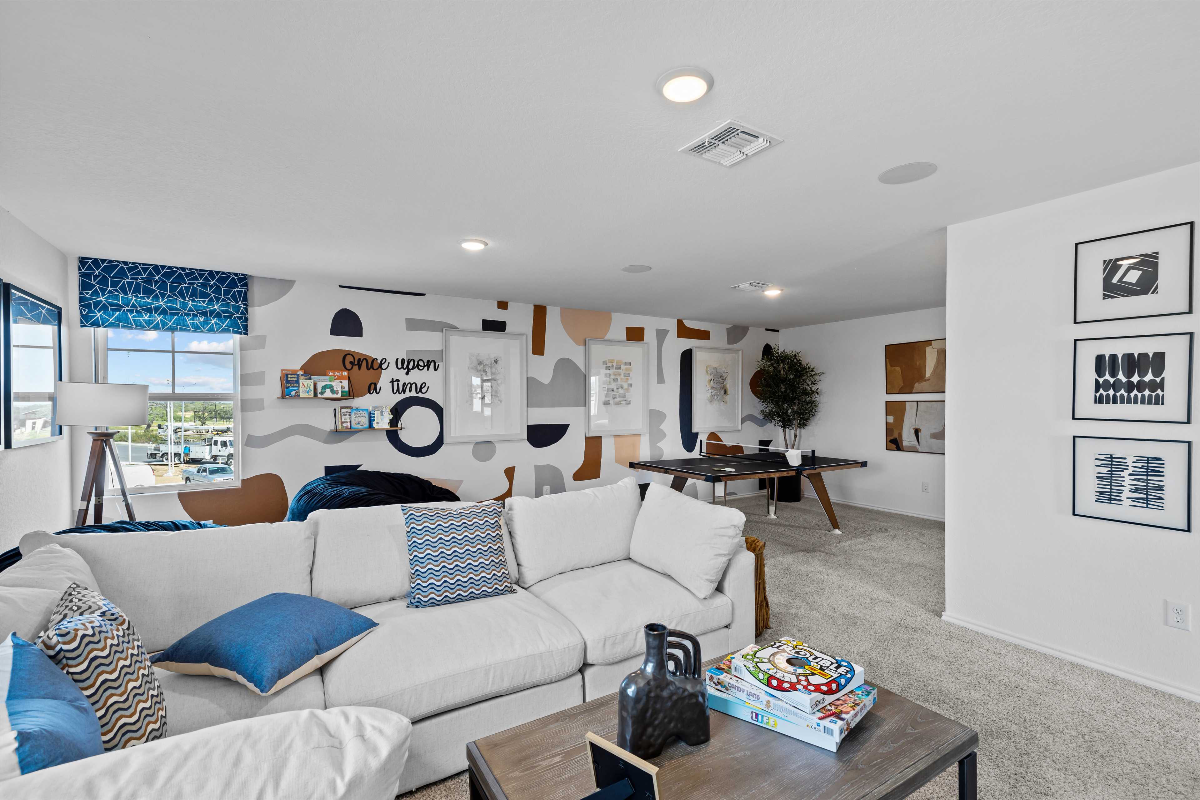 Cozy loft bonus room at Hickory Ridge in Elmendorf Texas by Davidson Homes featuring white sectional sofa blue pillows abstract wall art and coffee table
