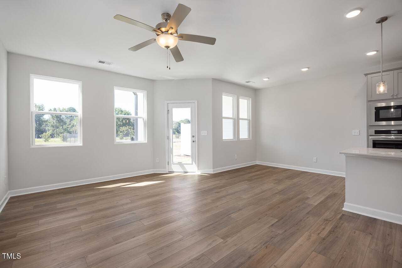 Bright open-concept living area with hardwood floors, ceiling fan, large windows, and kitchen island in The Graham Interior, Fuquay-Varina, NC