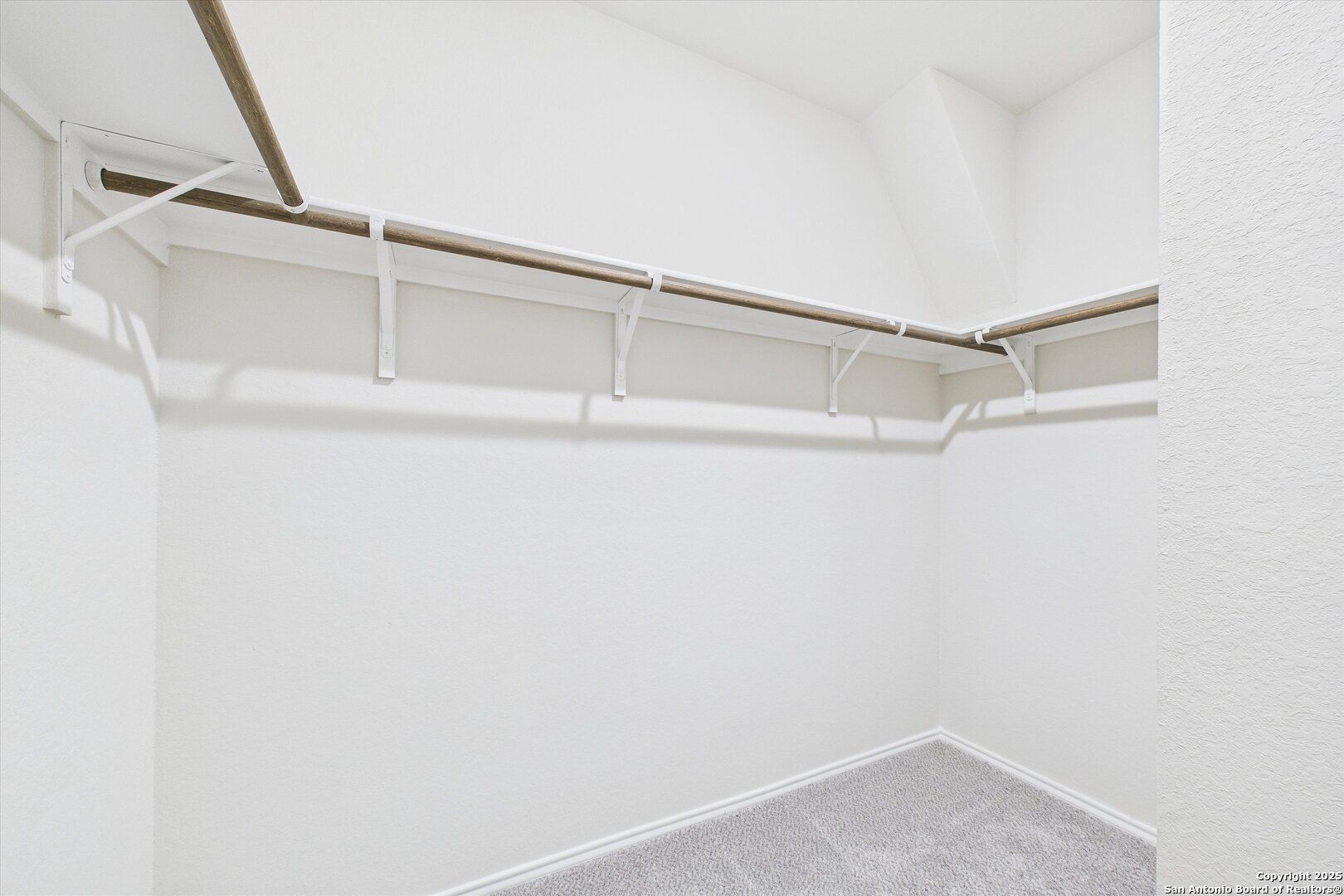 Spacious walk-in closet with wooden rods, white walls, and gray carpet in Davidson Homes The Asheville K, Bricewood, San Antonio