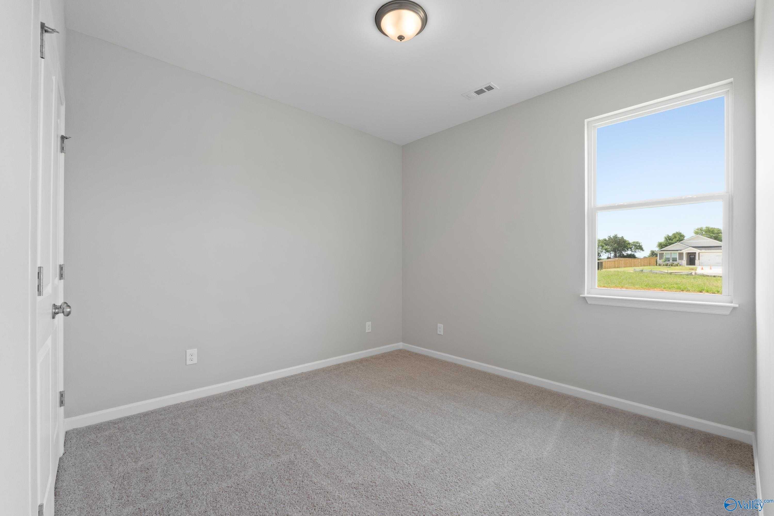 Empty bedroom with light gray walls, carpeted floor, and window overlooking fields in Evermore Homes The Aurora, Harvest, Alabama