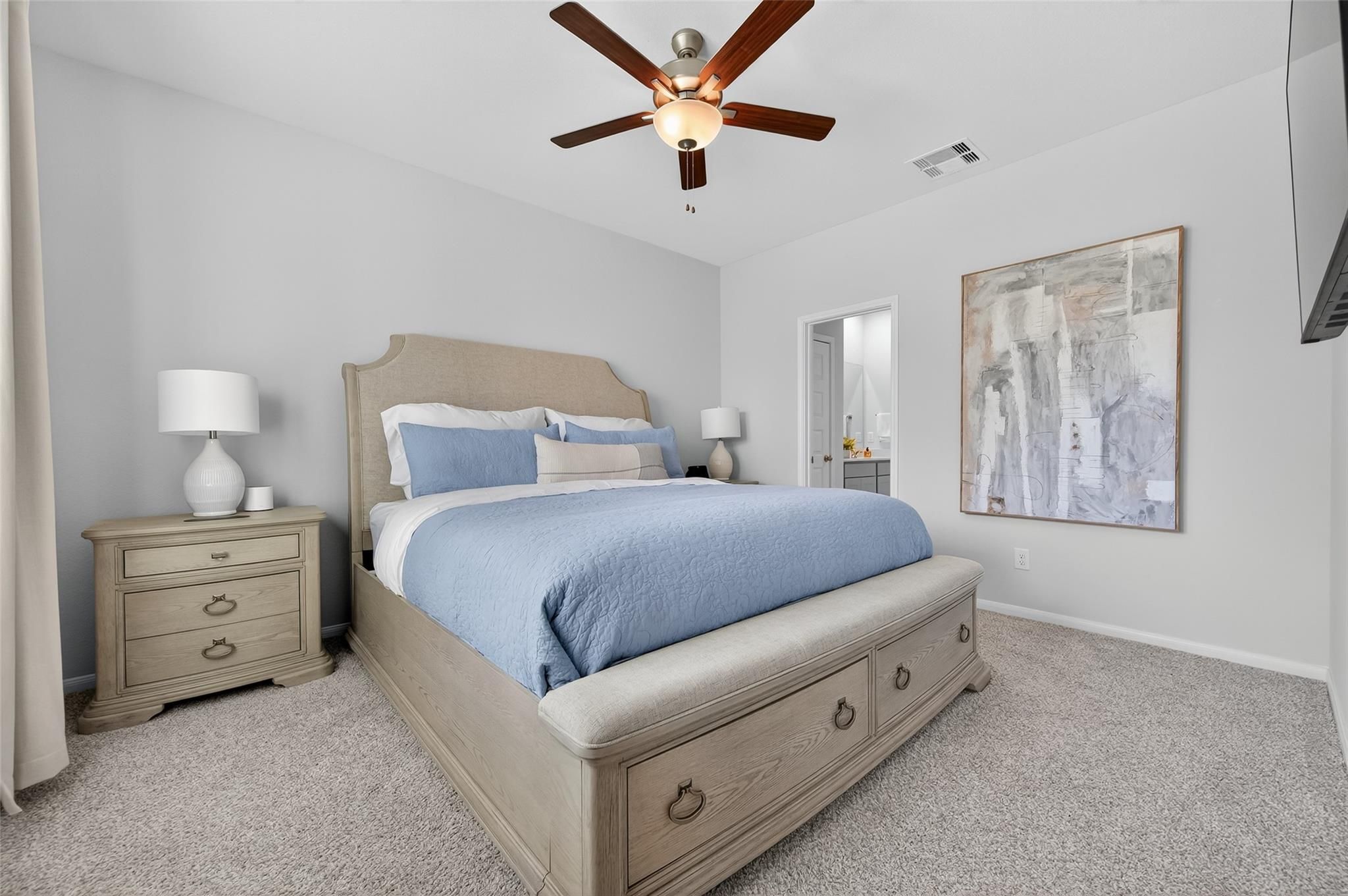 Cozy bedroom with beige upholstered bed, blue bedding, ceiling fan, wall art, and en-suite door in Davidson Homes The Brazos E, Magnolia, Texas