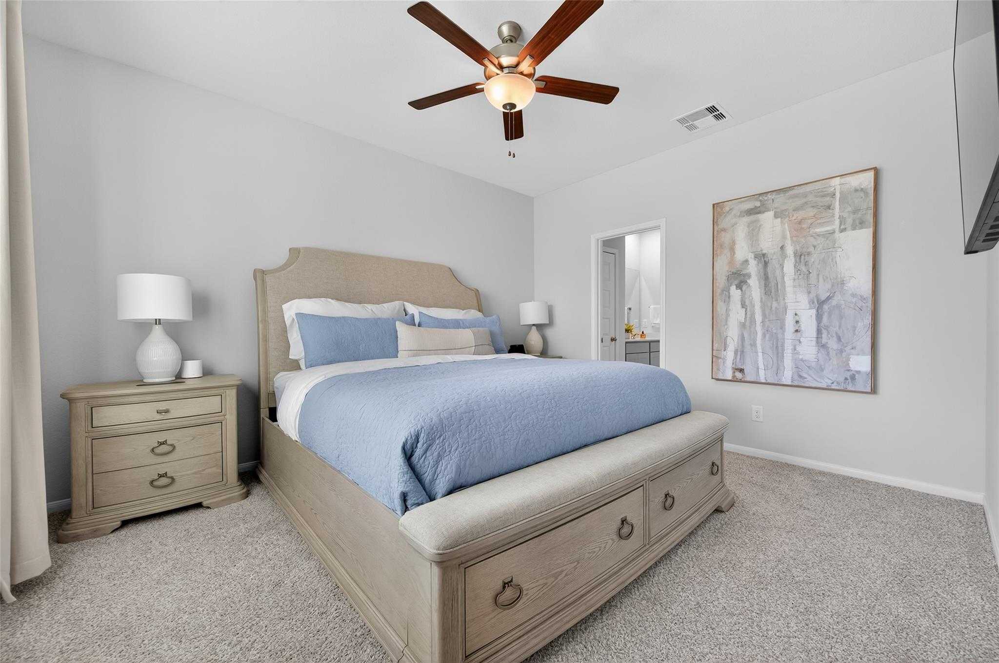 Cozy bedroom with beige upholstered bed, blue bedding, ceiling fan, wall art, and en-suite door in Davidson Homes The Brazos E, Magnolia, Texas
