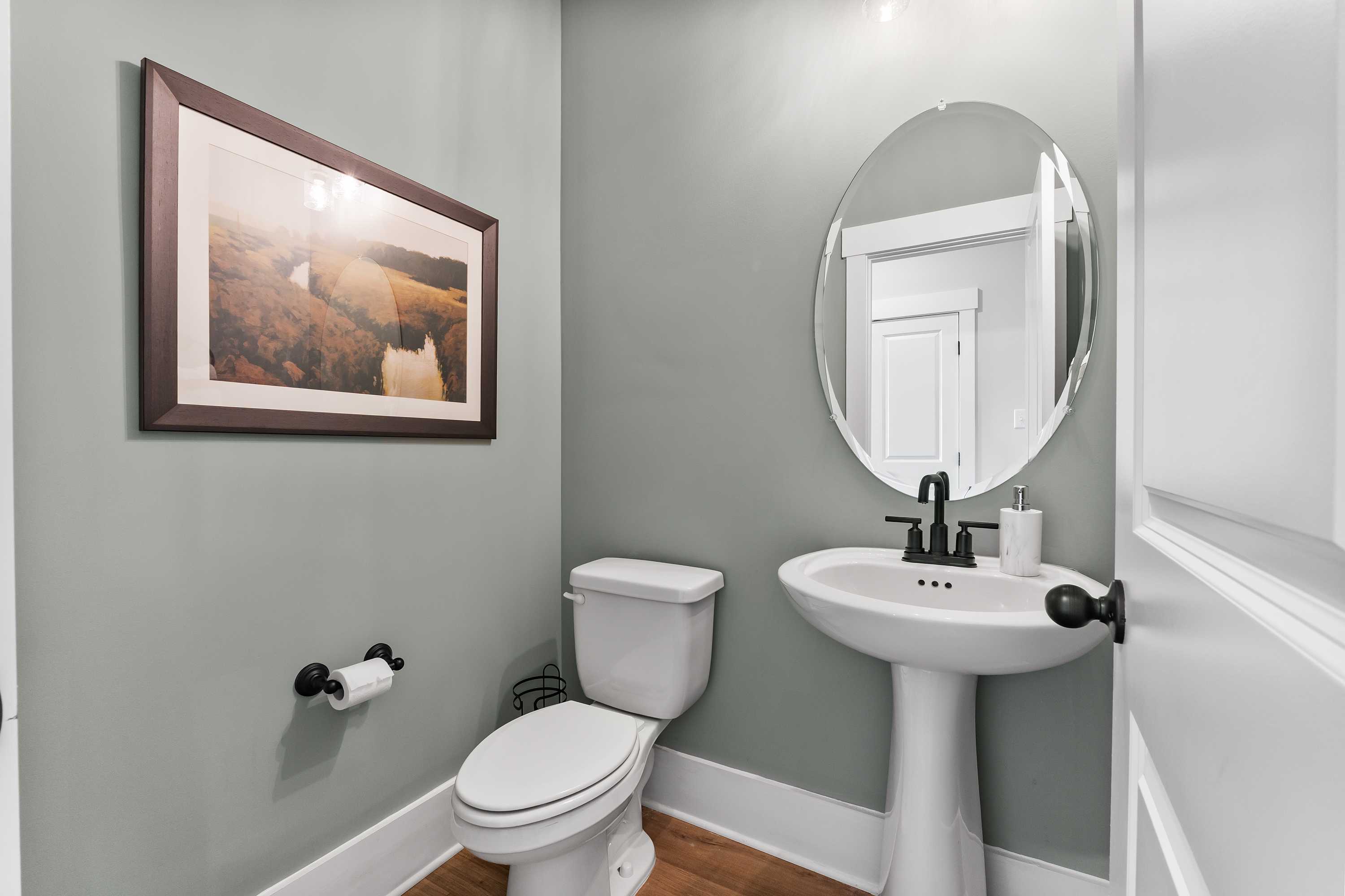 Cozy powder room at River Road Estates in Decatur AL with sage green walls, pedestal sink, oval mirror, and rustic landscape artwork