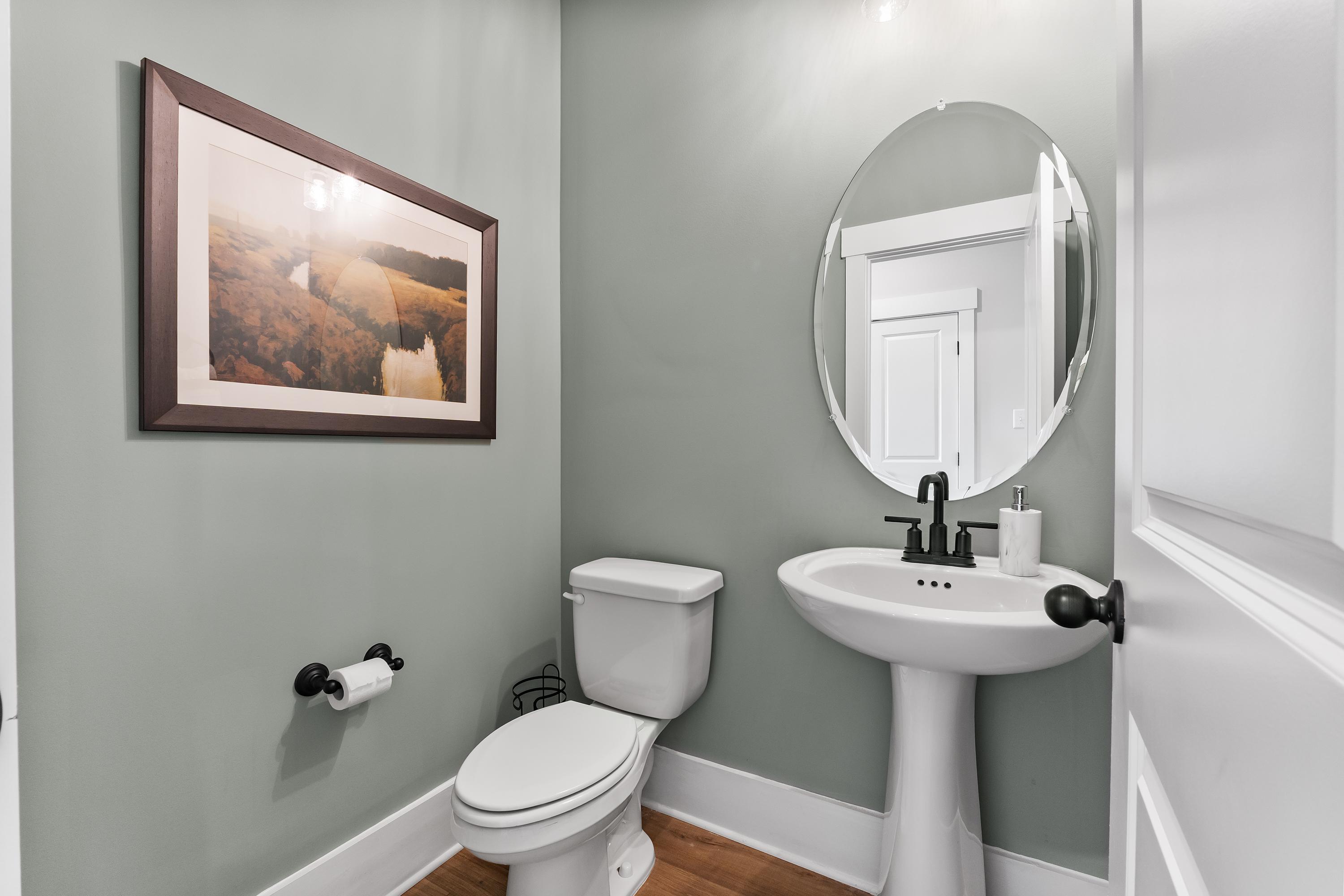 Cozy powder room at River Road Estates in Decatur AL with sage green walls, pedestal sink, oval mirror, and rustic landscape artwork