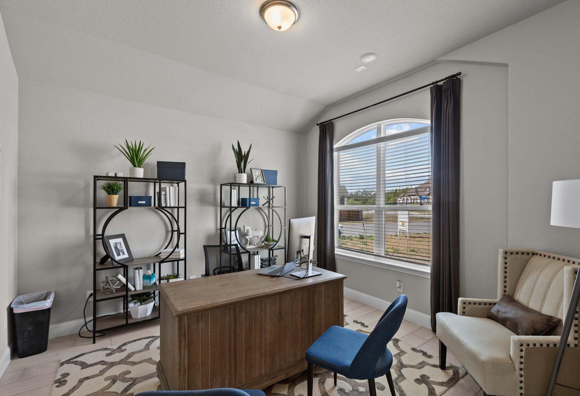 Modern home office in The Garner A featuring wooden desk, blue chair, bookshelves with plants, arched window, and neutral tones