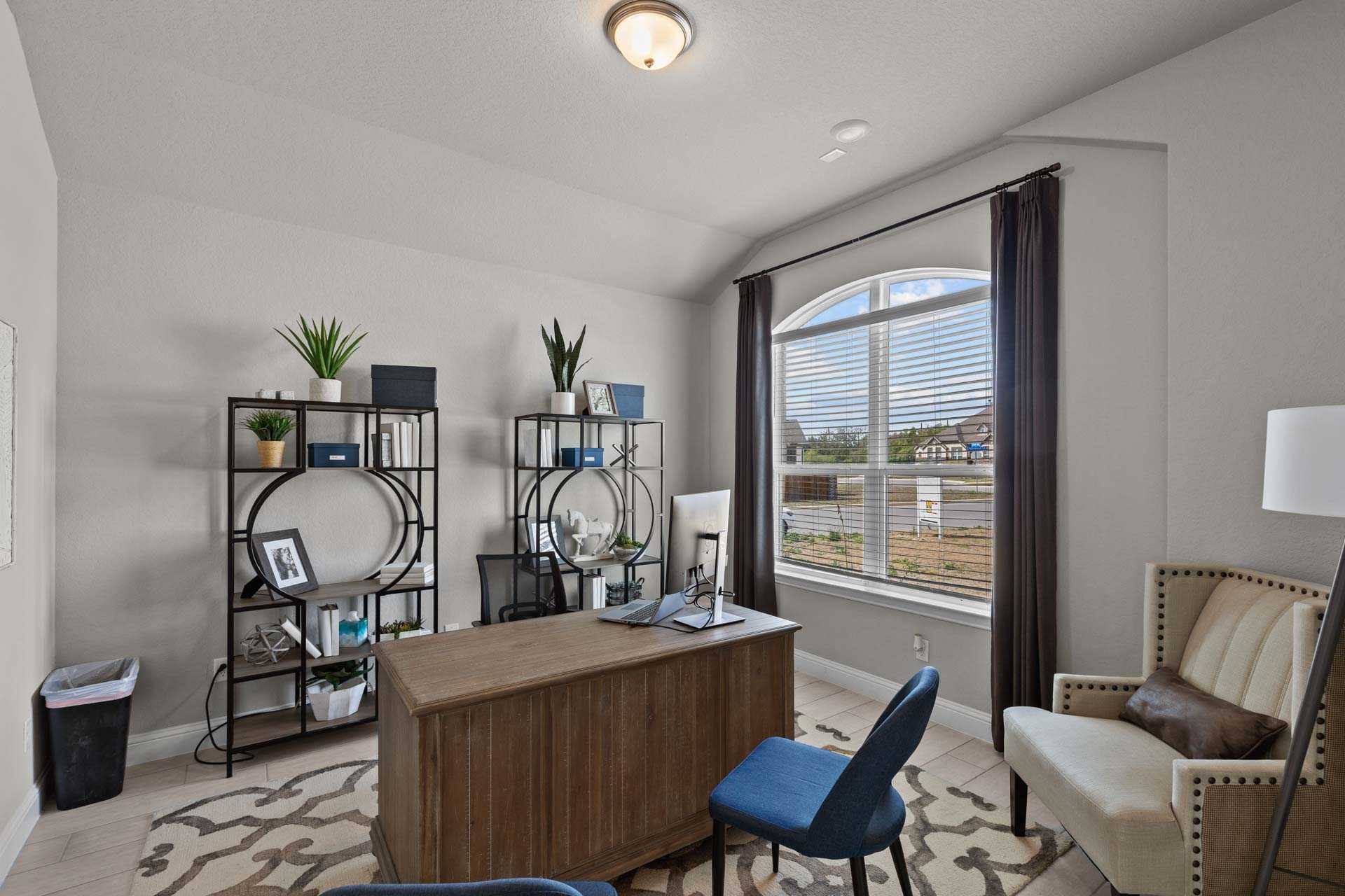 Modern home office in The Garner A featuring wooden desk, blue chair, bookshelves with plants, arched window, and neutral tones