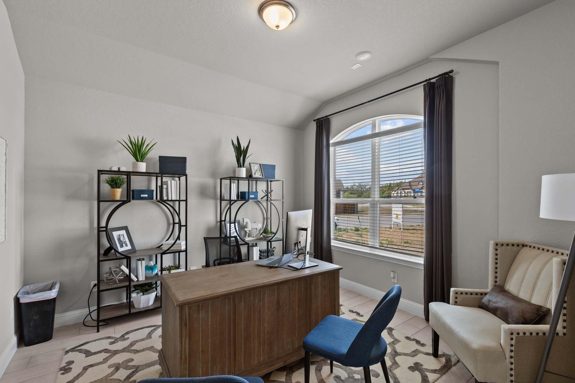 Modern home office in The Garner A featuring wooden desk, blue chair, bookshelves with plants, arched window, and neutral tones