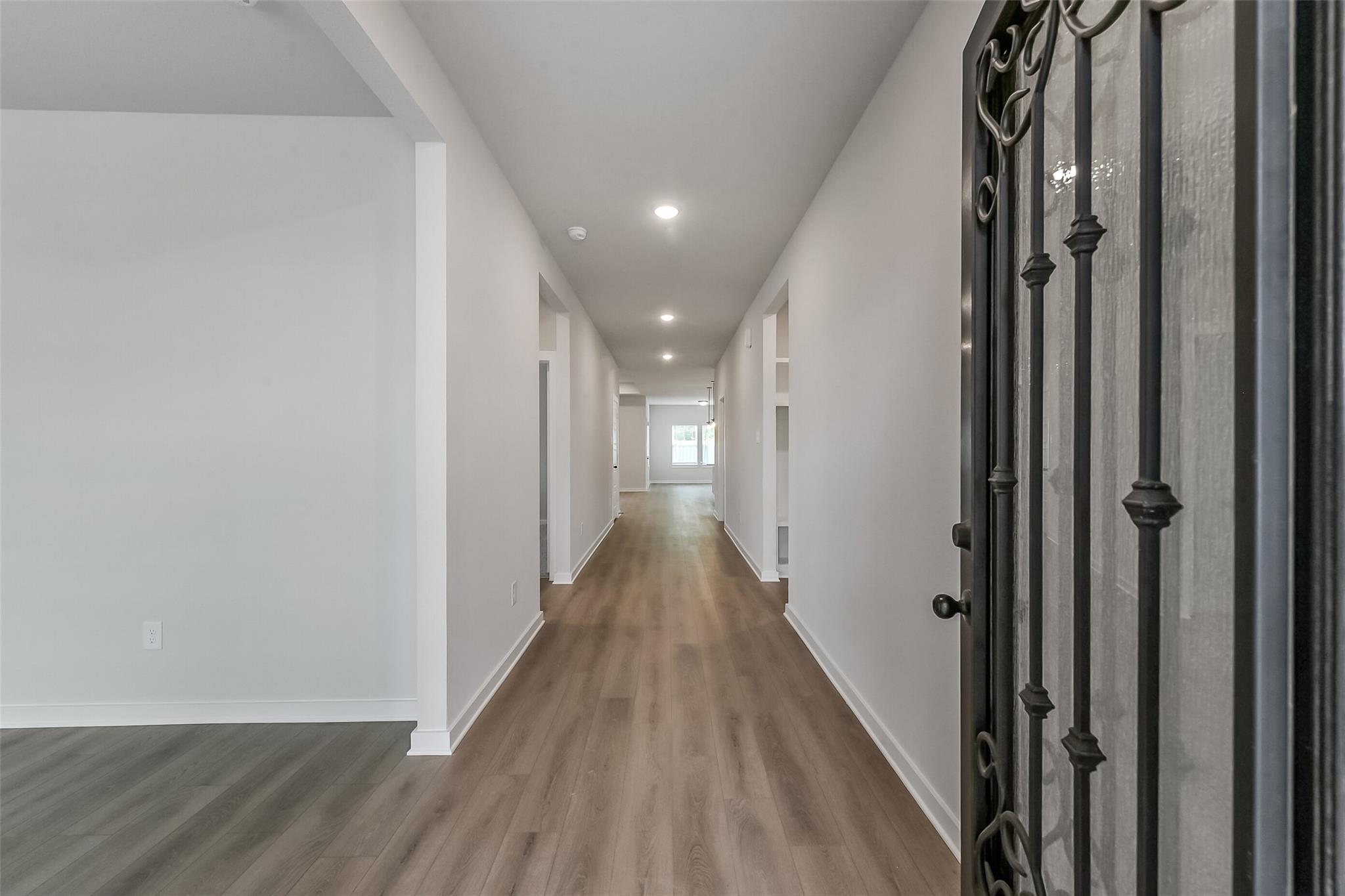 Elegant hallway with light wood flooring, white walls, recessed lights, and ornate black metal door in The Everett C by Davidson Homes, Crosby, TX