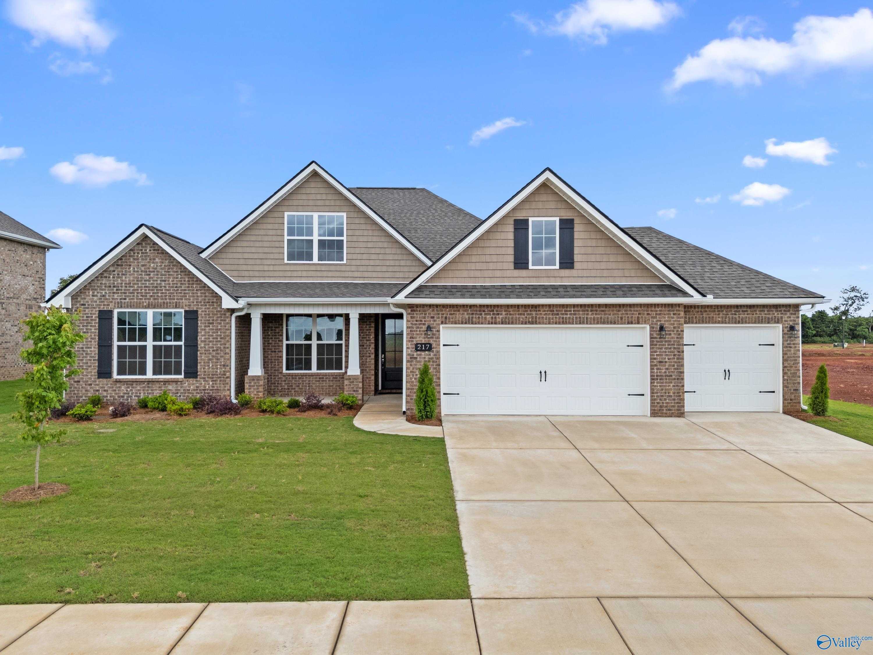 Modern tan brick single-story home with 3-car garage, covered porch, and lush lawn in Kendall Farms, Toney, Alabama