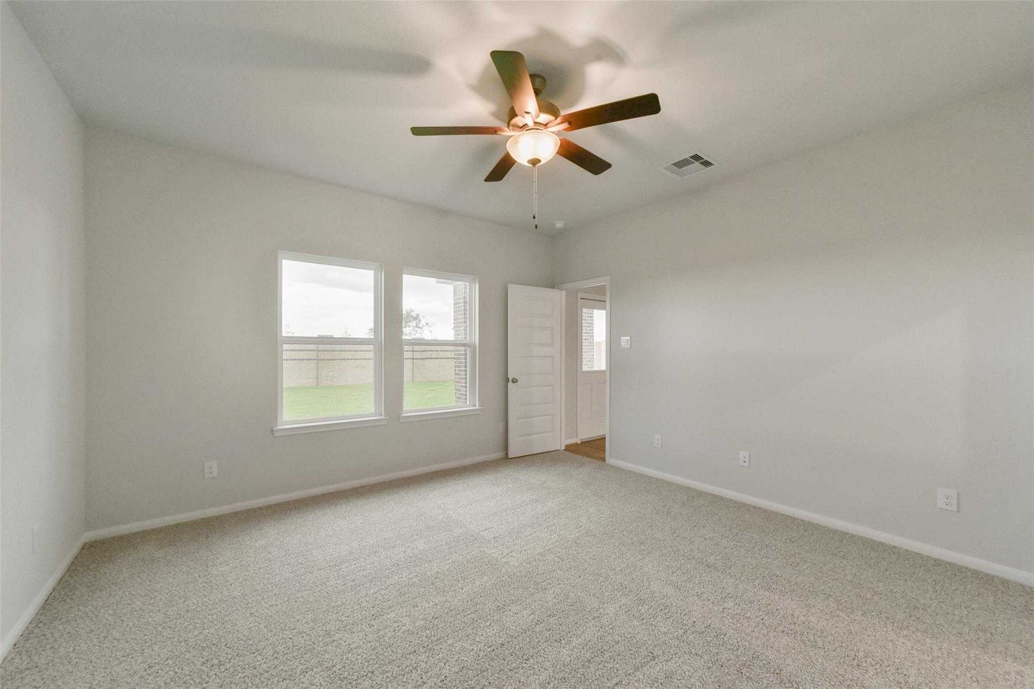 Spacious bedroom with gray walls, beige carpet, ceiling fan, and large windows overlooking yard in Davidson Homes The San Marcos E, Beasley, Texas