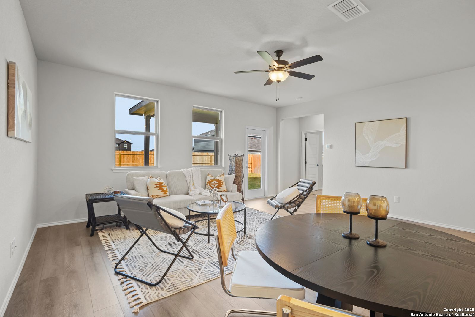 Cozy living-dining area with gray sofa, round wood table, ceiling fan, and backyard view through large windows in Davidson Homes The Douglas C, Seguin Texas