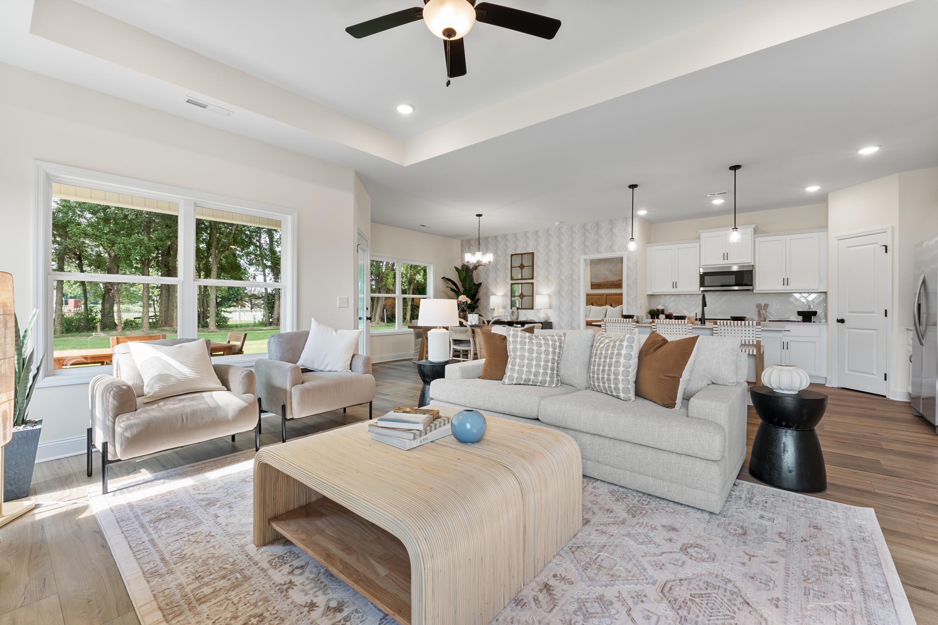 Open-concept living room and kitchen at Lynn Meadows in Meridianville AL by Davidson Homes with beige sofa, wooden coffee table, large windows, white cabinets
