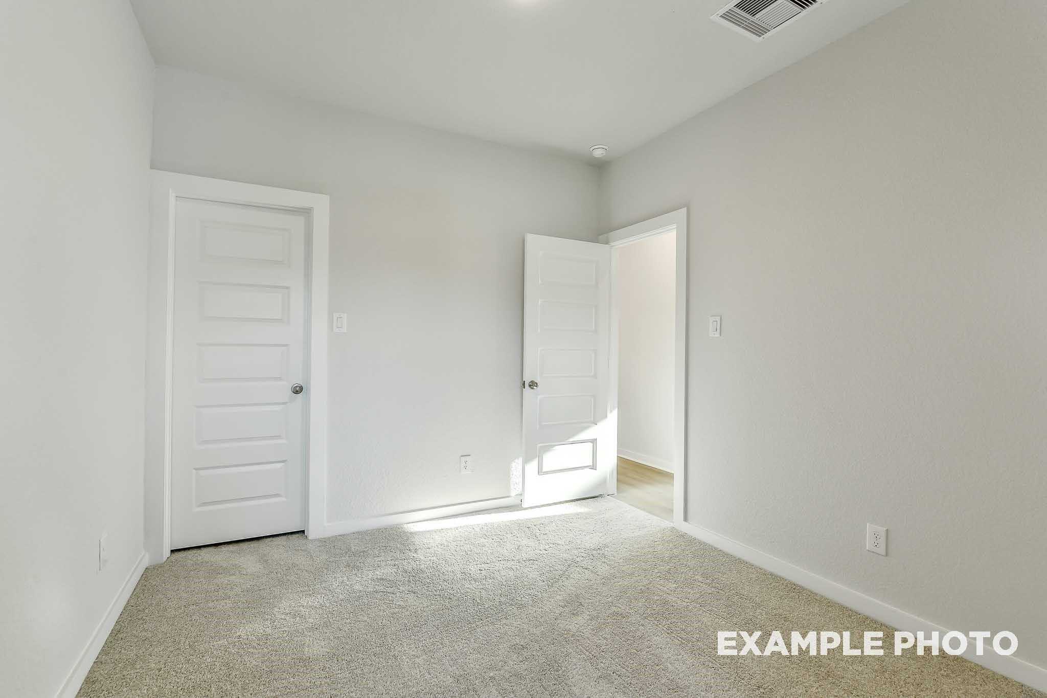 Spacious empty bedroom with light gray walls, beige carpet, and open white door in Davidson Homes The Colorado F, Conroe, Texas