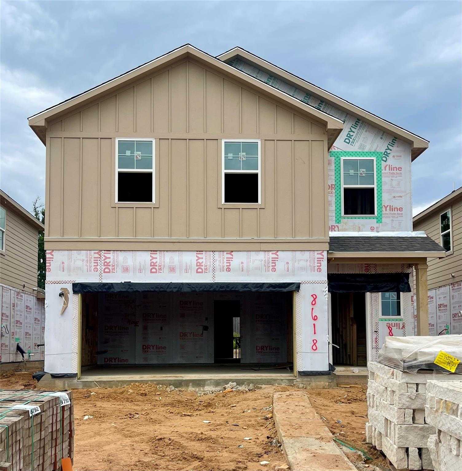 Two-story beige Davidson Homes The Blanco E with open 2-car garage, house wrap, and stacked bricks in Lakes at Black Oak, Magnolia, Texas