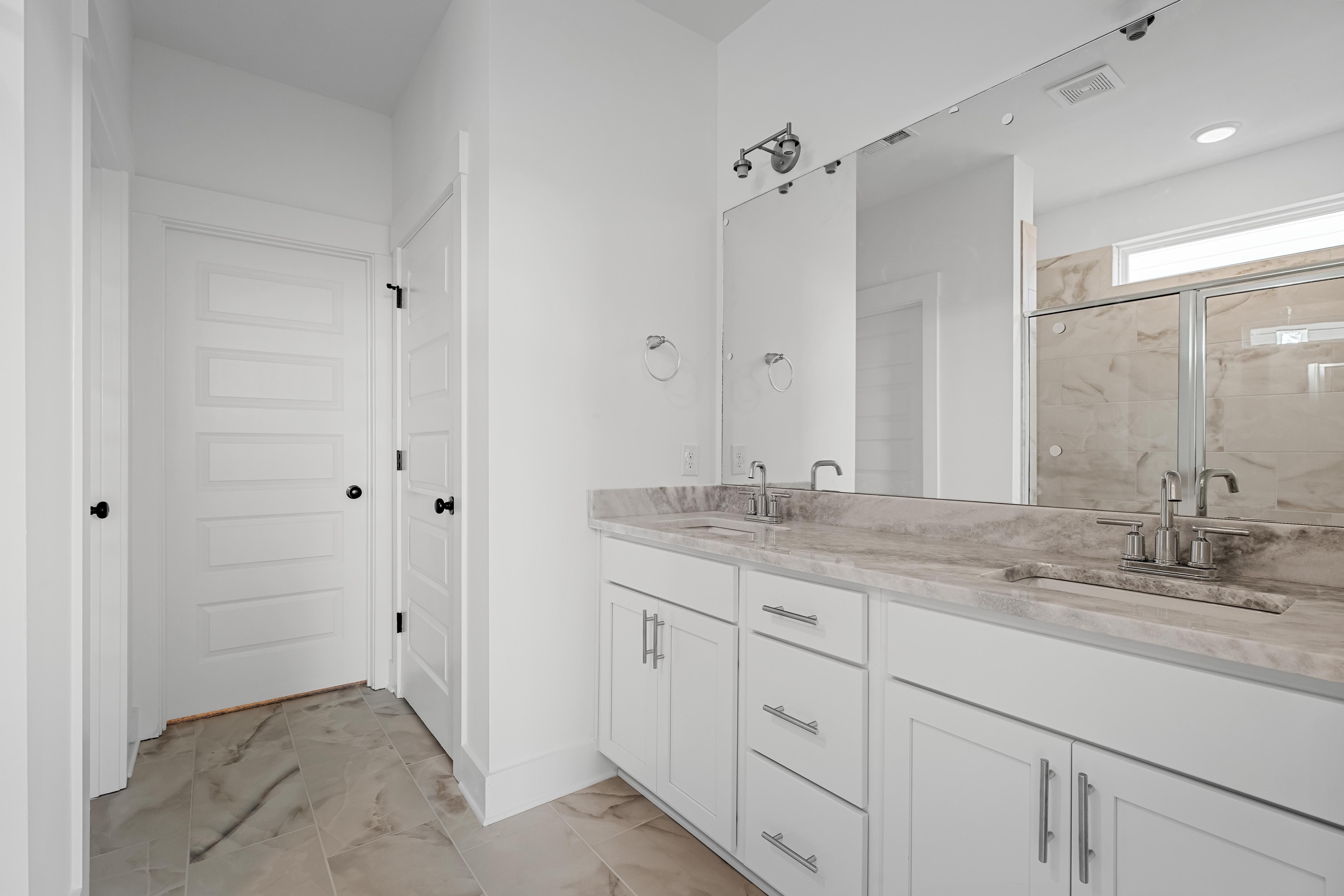 Modern primary bathroom in The Cedar D featuring a dual-sink vanity with marble countertops and a glass-enclosed shower.