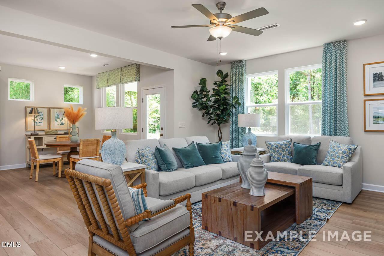 Cozy open-concept living room with gray sectional sofa, rattan chairs, dining table, and large windows in Davidson Homes Willow B, Wake Forest, NC
