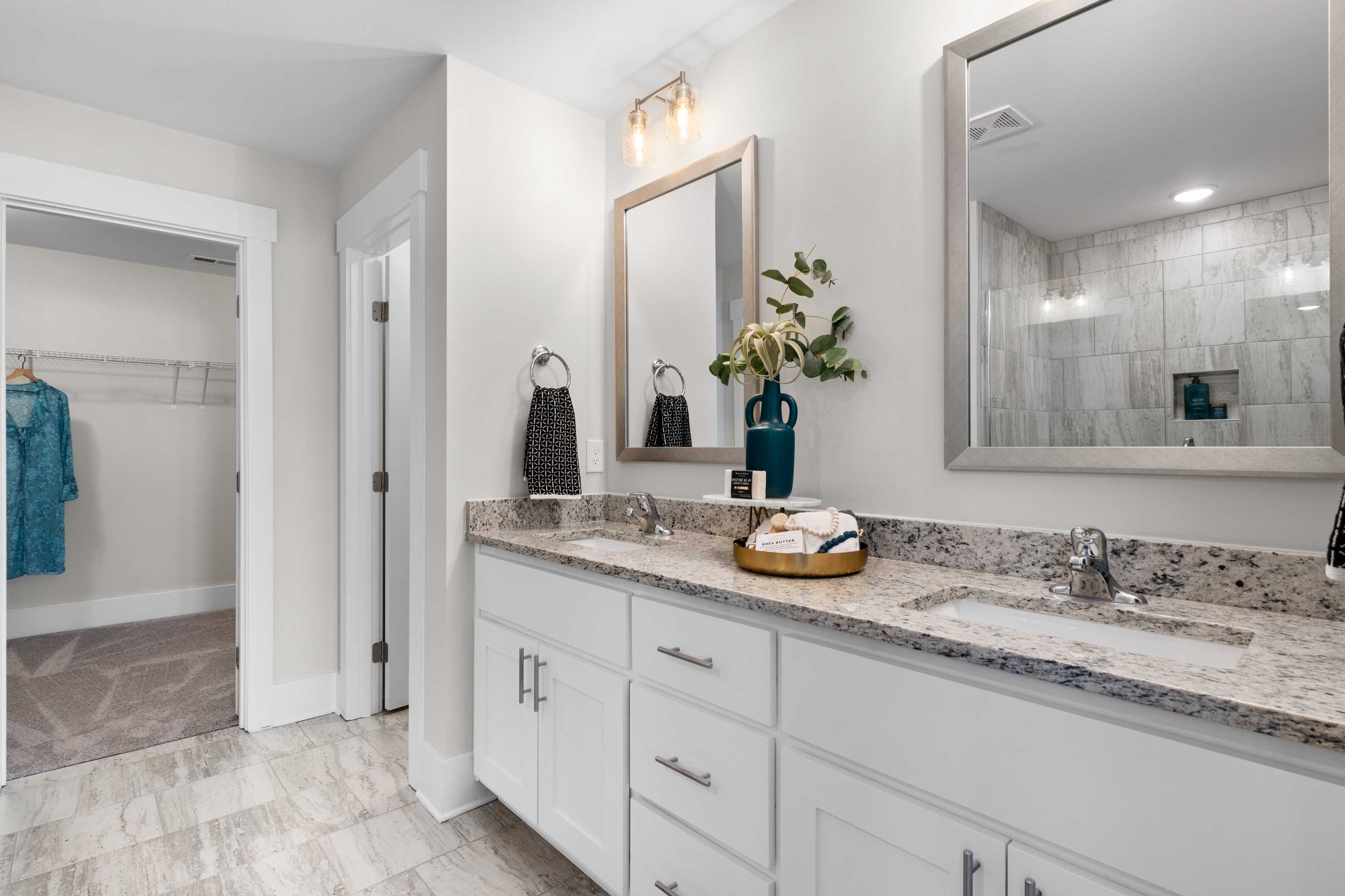 Spacious master bathroom in The Chelsea B with double white vanity, granite counters, frameless mirrors, tiled walk-in shower, and adjacent walk-in closet