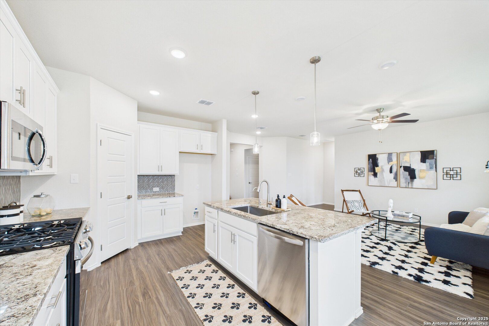 Modern open-concept kitchen with granite island, white cabinets, stainless appliances, and adjacent living area in The Asheville K by Davidson Homes, San Antonio