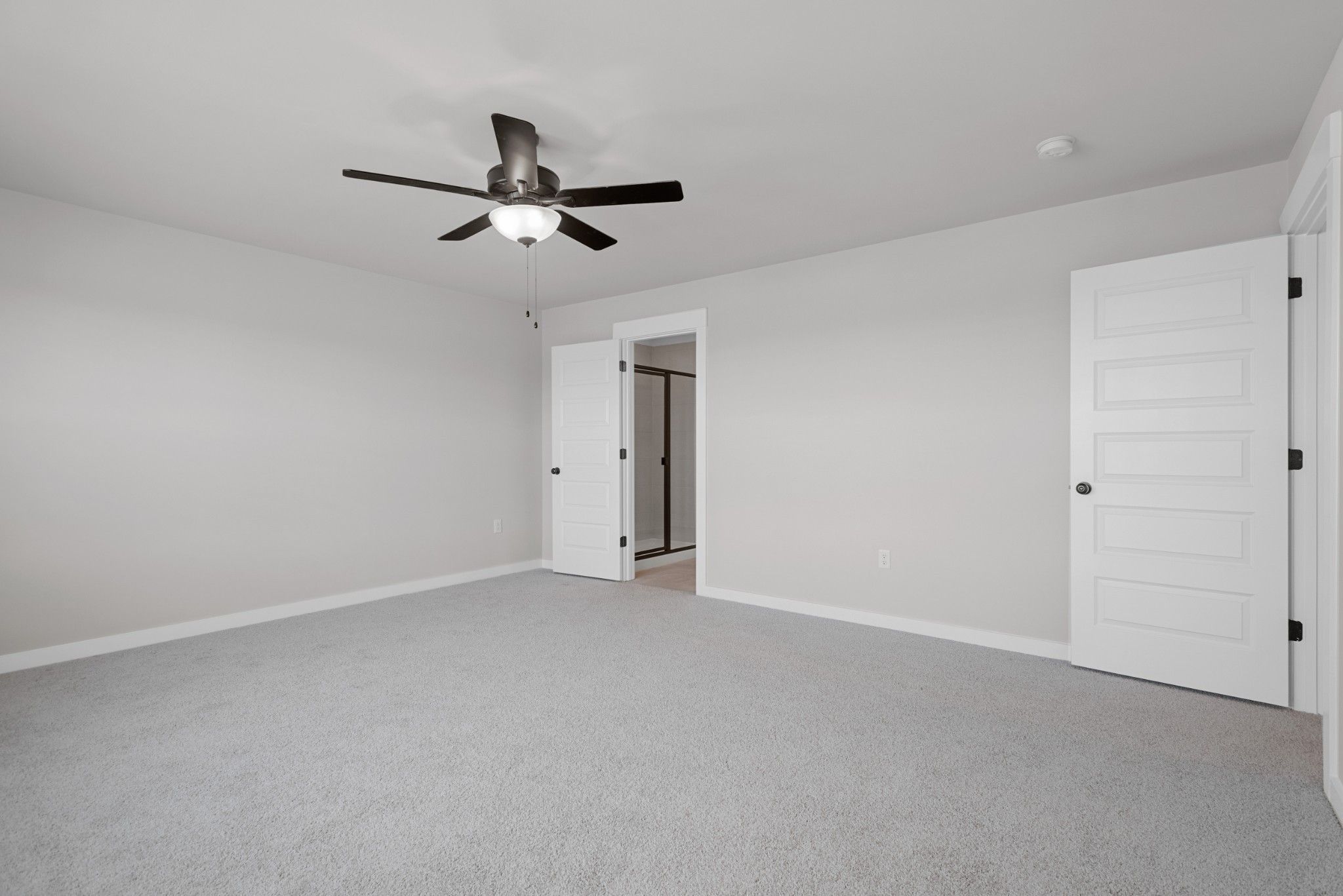 Spacious bedroom with grey carpeting, ceiling fan, and en-suite bathroom door in The Willow B plan at Calista Farms.