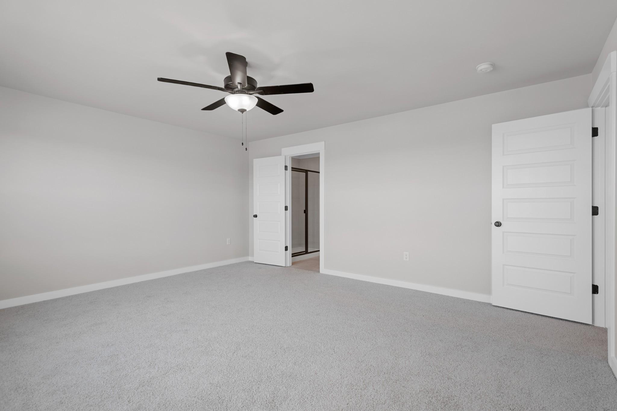 Spacious bedroom with grey carpeting, ceiling fan, and en-suite bathroom door in The Willow B plan at Calista Farms.