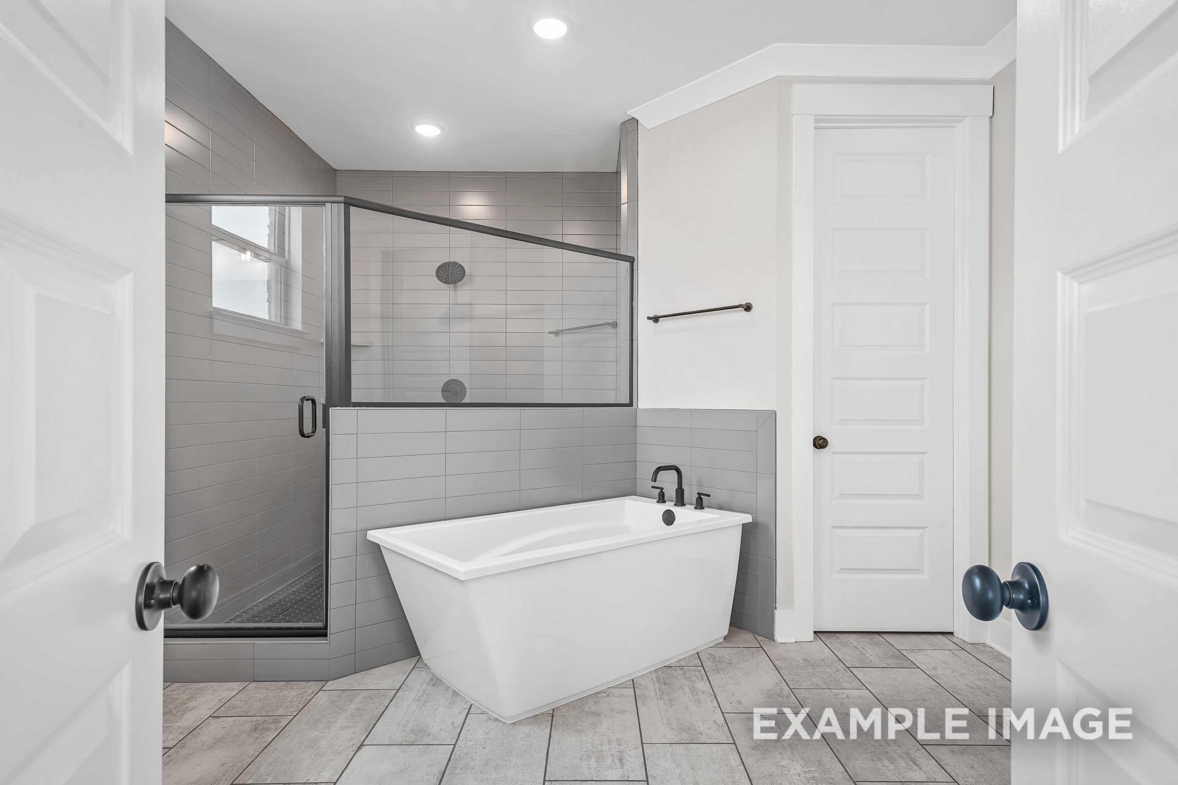 Luxury master bathroom in The Hawkins showcasing freestanding white tub, glass shower, gray tiles, and double doors