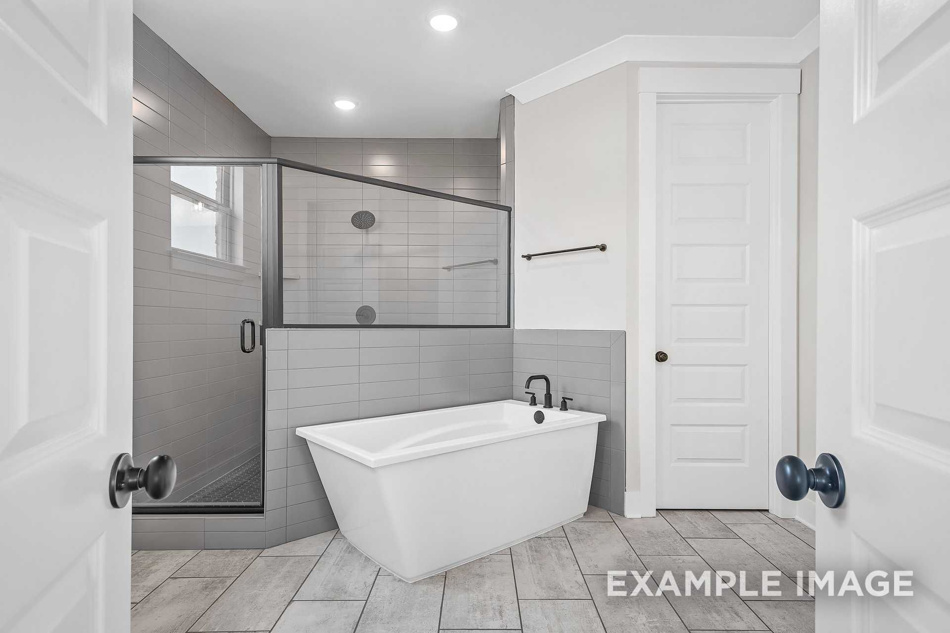 Luxury master bathroom in The Hawkins showcasing freestanding white tub, glass shower, gray tiles, and double doors