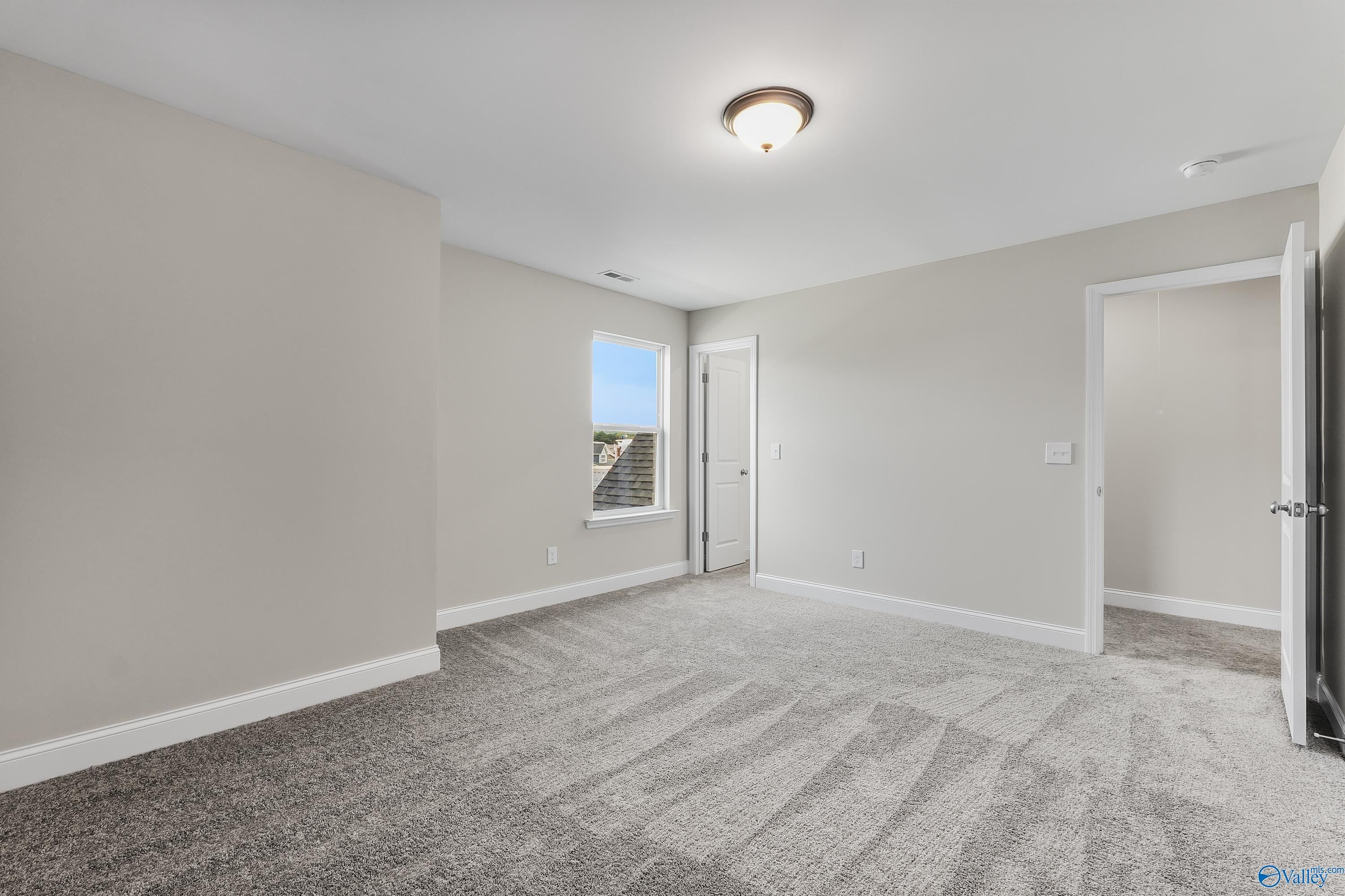 Spacious empty bedroom with neutral beige walls, plush carpet, large window, and adjacent door in Evermore Homes The Haven, Owens Cross Roads, AL