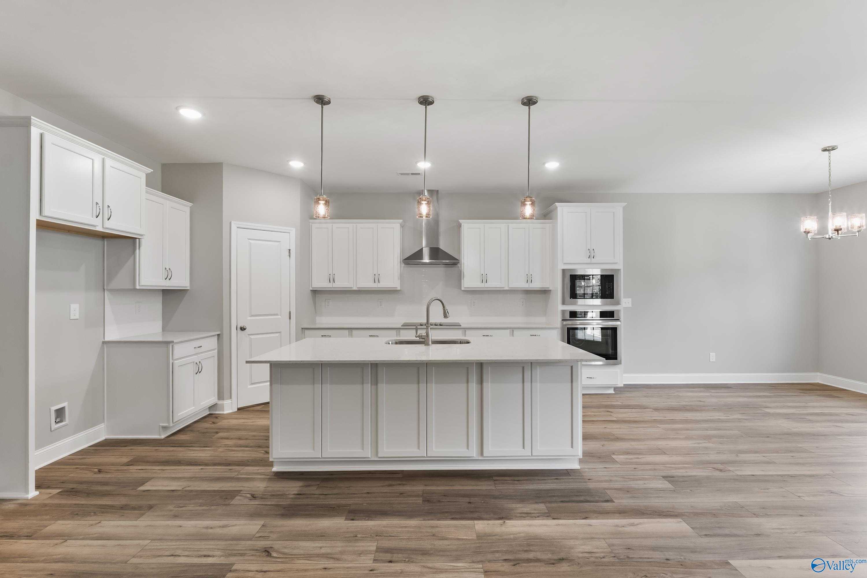 Modern white kitchen island with sink, stainless appliances, pendant lights, and hardwood floors in Davidson Homes The Rockford, Harvest, AL