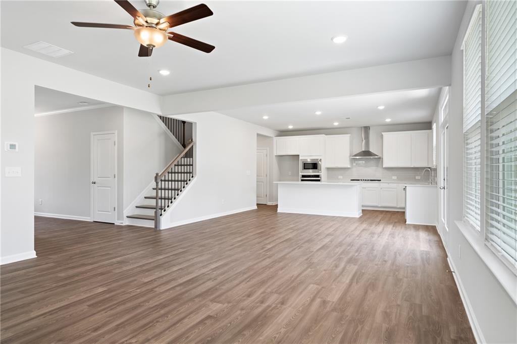 Open-concept great room with hardwood floors, white kitchen island, staircase, and ceiling fan in The Hickory A, Winder, GA