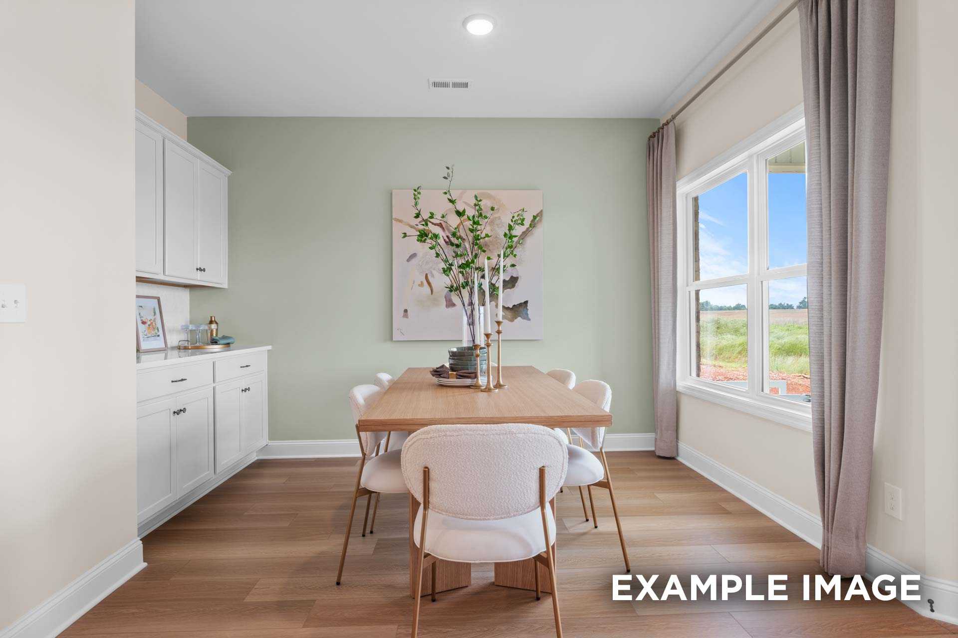 Spacious dining area in The Laurel N home with wooden table, white chairs, abstract sage wall art, and large windows overlooking fields