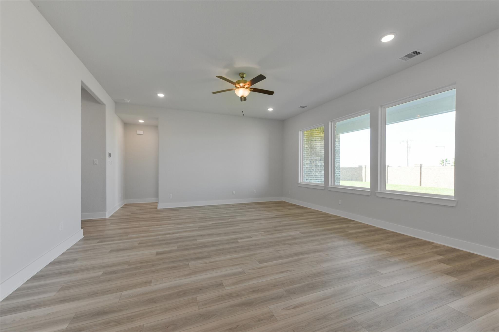 Spacious empty living room with light wood laminate flooring, ceiling fan, recessed lights, and large windows in Davidson Homes The Edward C, Lago Mar, Texas City