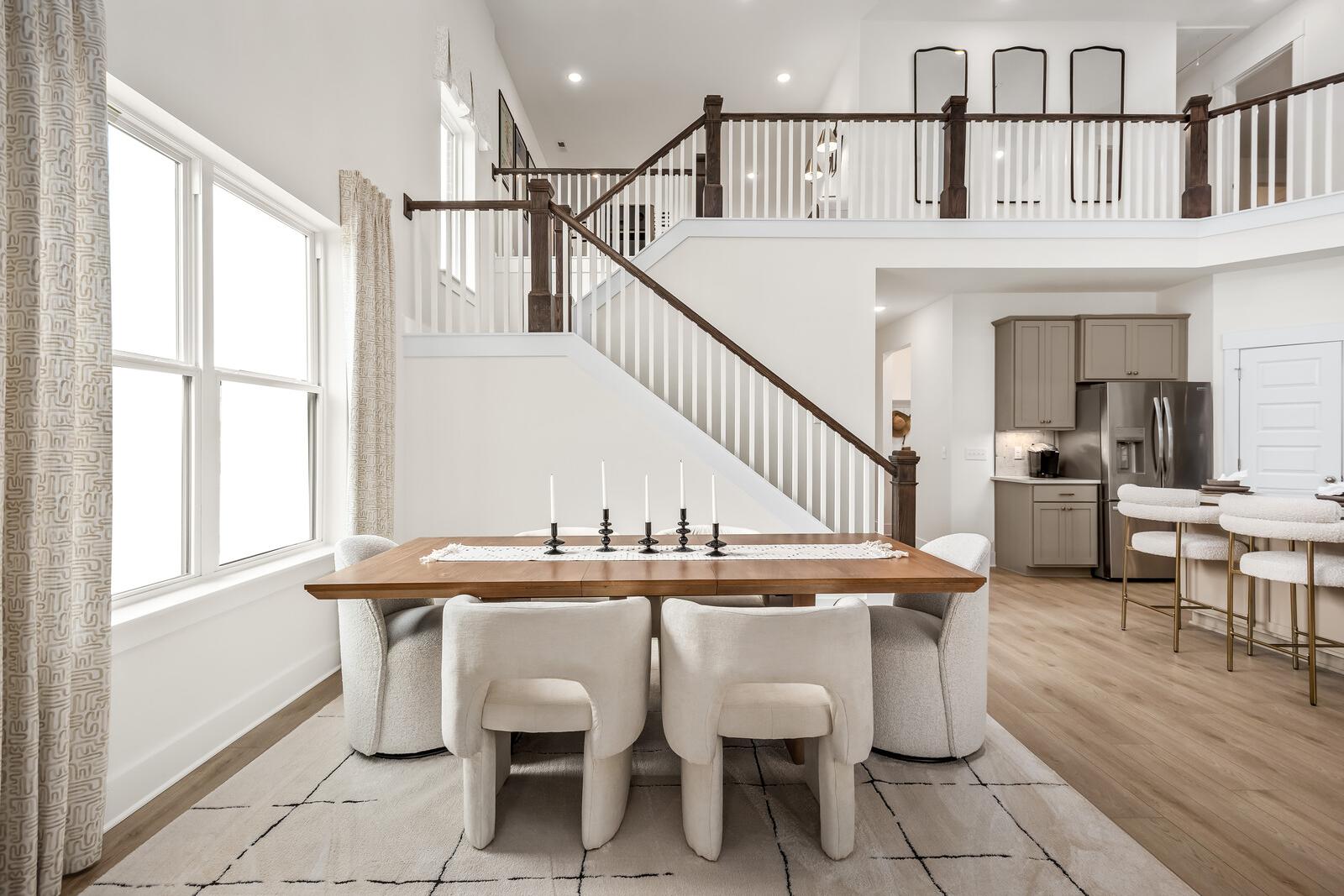 Open-concept dining room in The Ridgeport with wooden table, white upholstered chairs, grand wooden staircase to upper balcony, and adjacent kitchen
