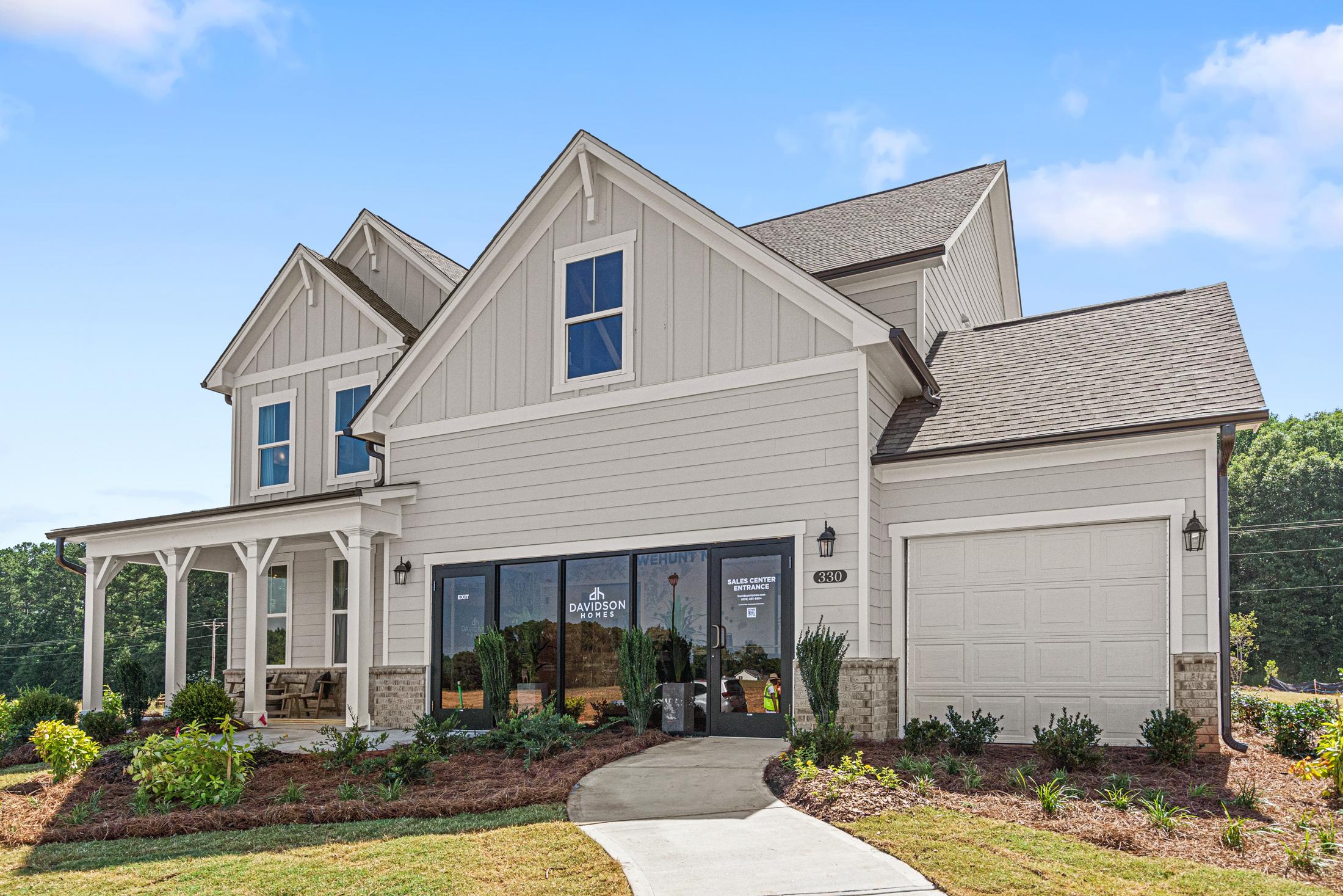 Two-story craftsman elevation of The Hickory B showcasing beige siding, covered porch, two-car garage, and lush landscaping in Hoschton