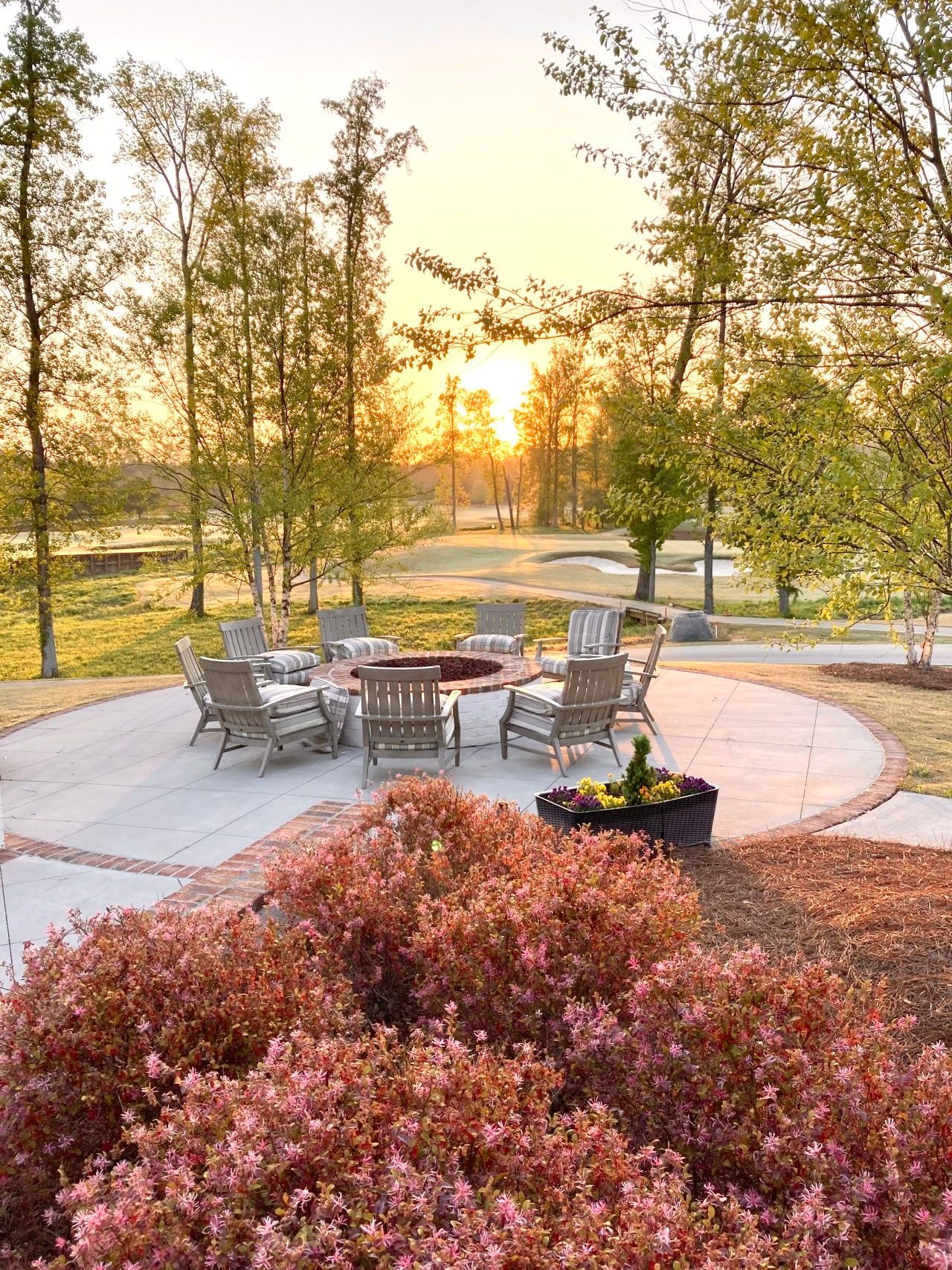 Cozy circular fire pit patio at Addison West in Holly Springs NC with Adirondack chairs, potted flowers and sunset trees