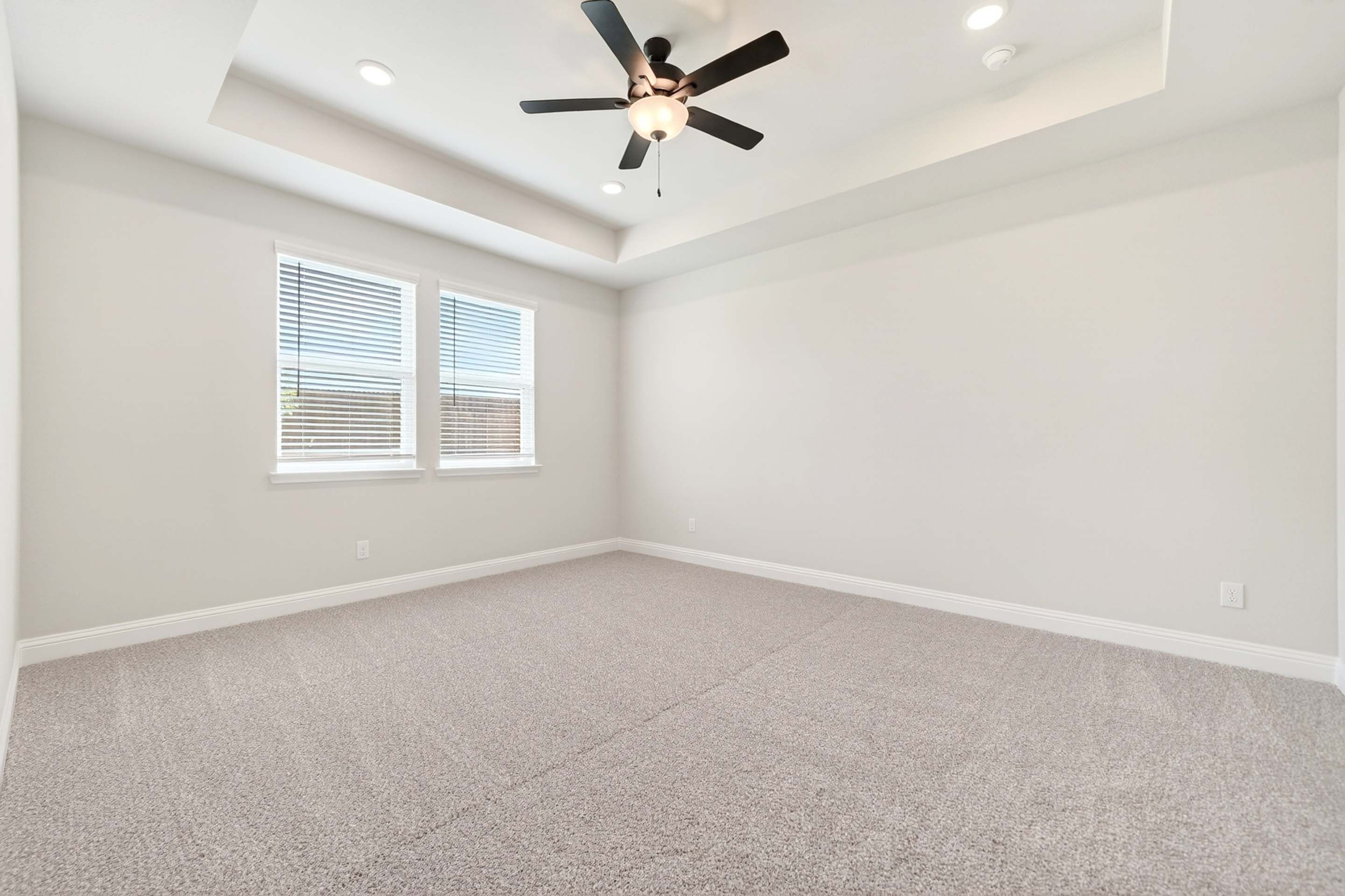 Spacious master bedroom in The Holly design featuring a tray ceiling, ceiling fan, and plush carpet in Royse City, Texas.