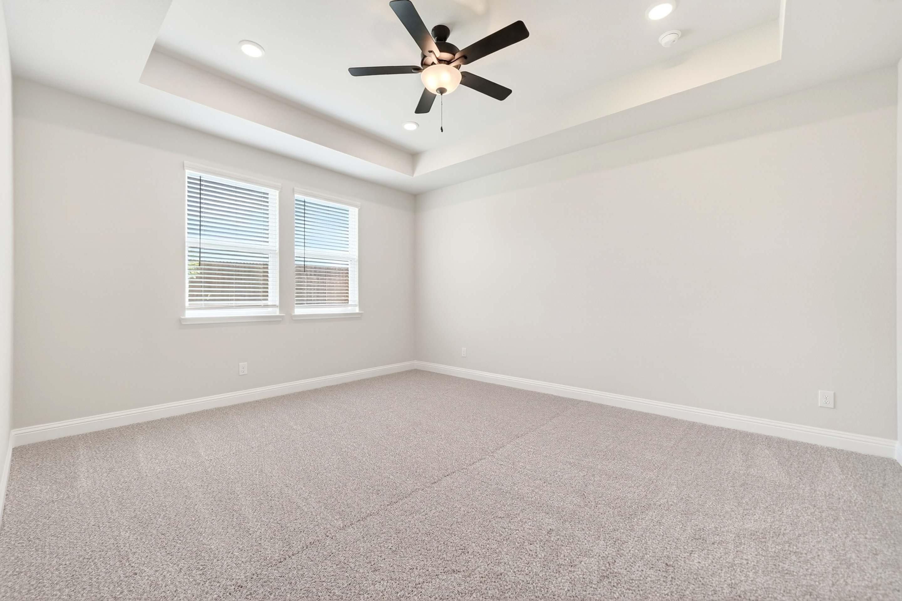 Spacious master bedroom in The Holly design featuring a tray ceiling, ceiling fan, and plush carpet in Royse City, Texas.