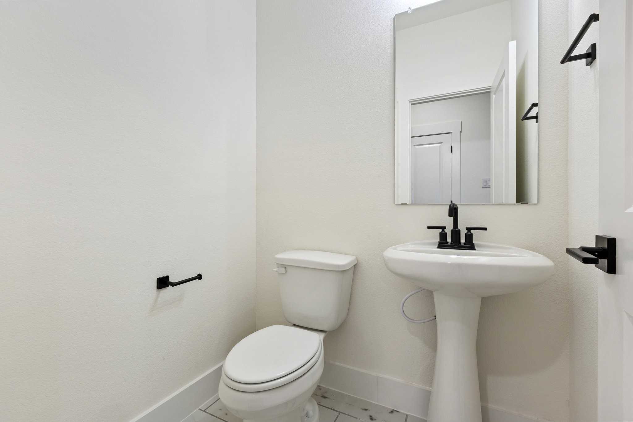 Elegant powder room featuring white pedestal sink, black faucet, toilet, and mirror in Davidson Homes The Rockford C, Josephine, Texas