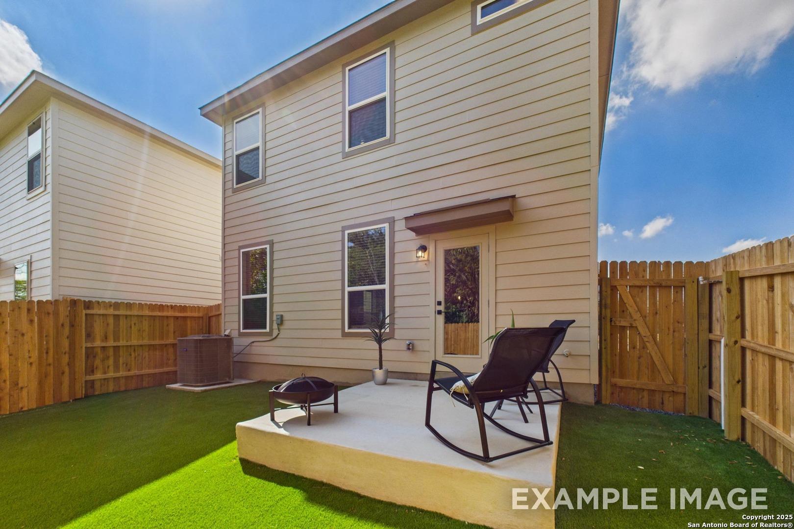 Two-story beige home exterior with covered porch, fenced backyard, green lawn, rocking chair and fire pit patio in Meadows at Oak Creek, San Antonio, Texas