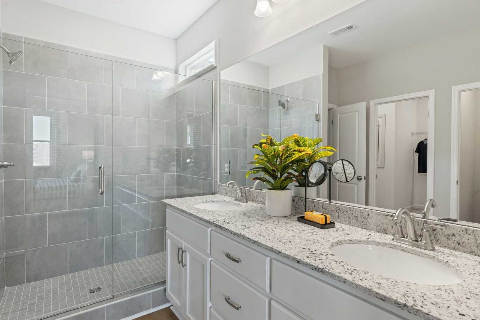 Spacious master bathroom in The Phoenix featuring frameless glass shower with gray tiles, double quartz vanity, and potted plants