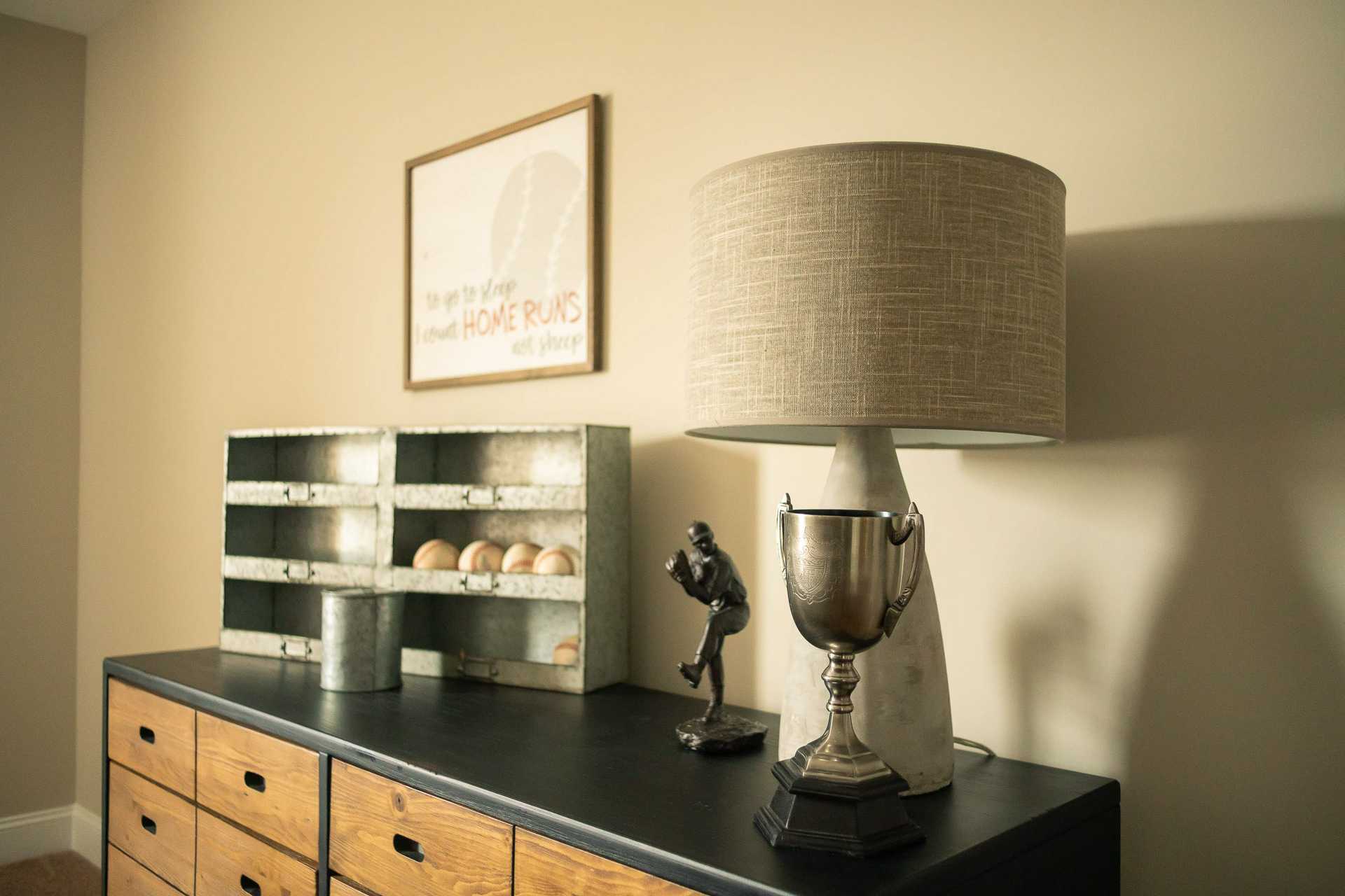 Rustic wooden dresser with galvanized bin of eggs, baseball trophy, figurine, and linen lamp in Summer Pointe Madison AL home