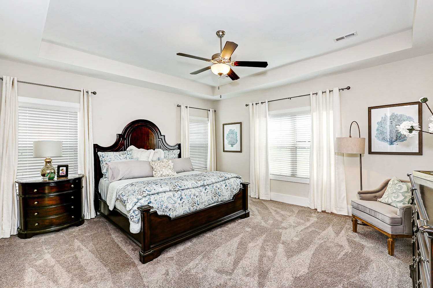 Spacious master bedroom in The Finleigh featuring king bed with ornate wood headboard, ceiling fan, large windows, and plush carpet
