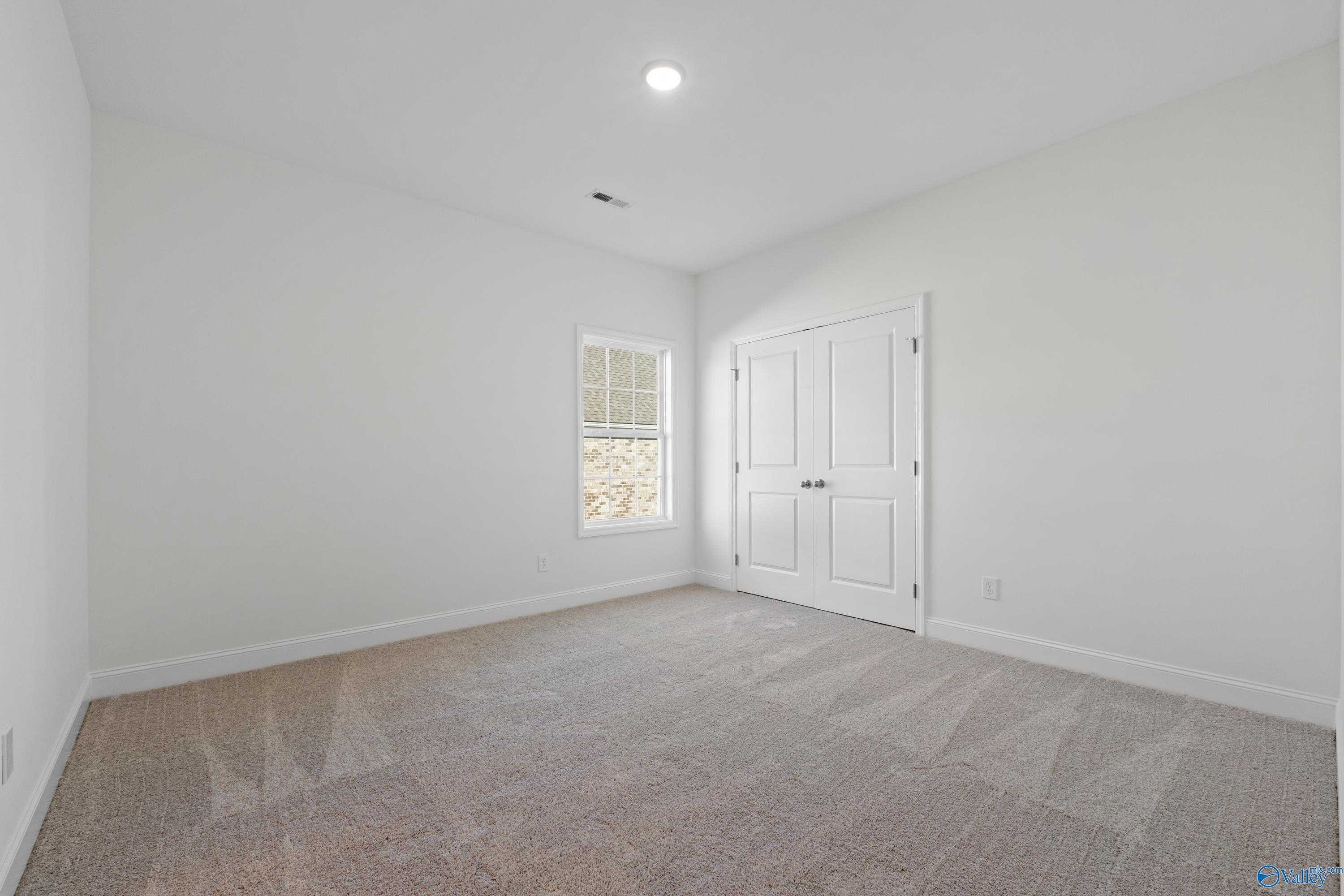 Spacious empty bedroom with white walls, beige carpet, large window, and double closet doors in Davidson Homes The Oxford, Harvest, AL