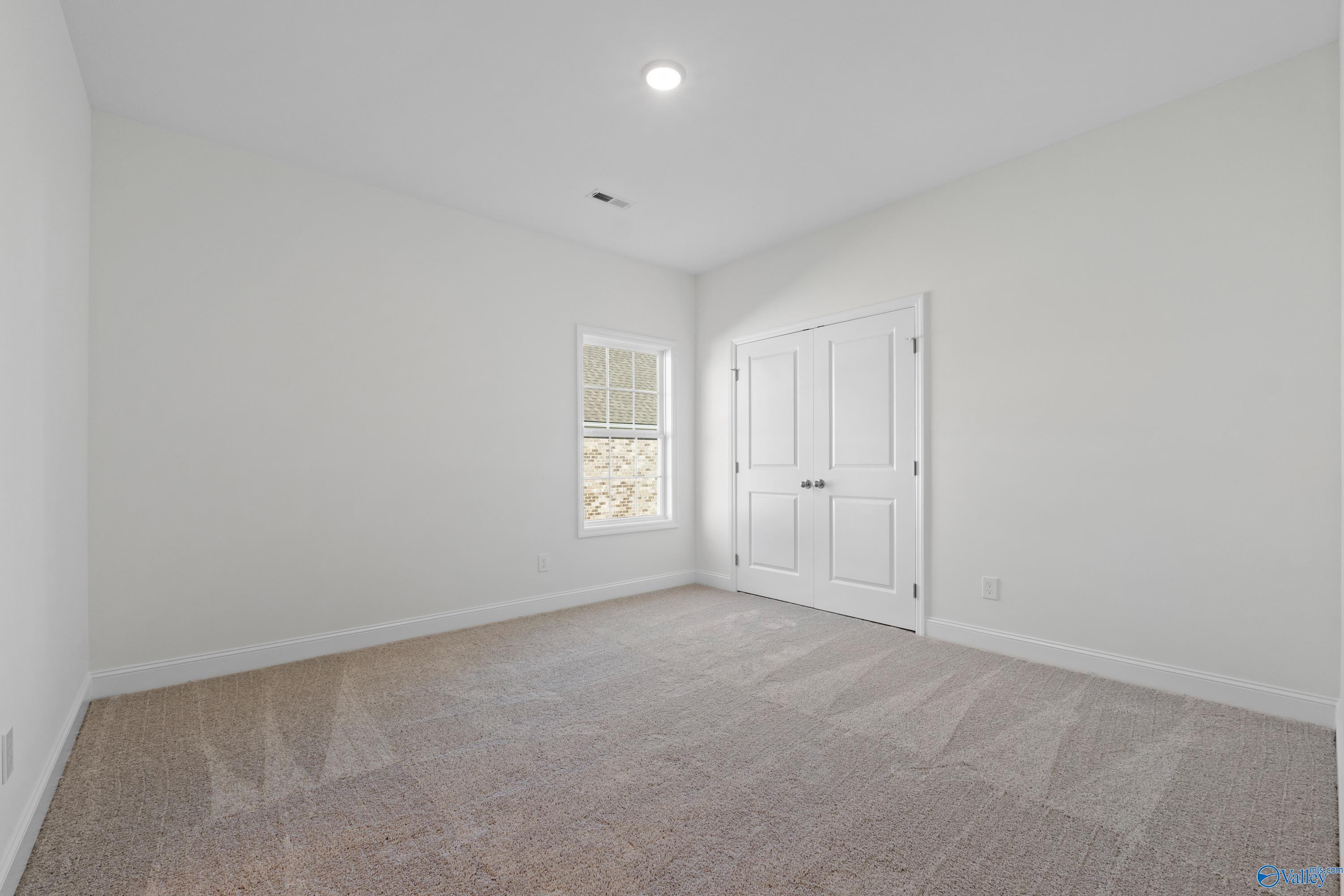 Spacious empty bedroom with white walls, beige carpet, large window, and double closet doors in Davidson Homes The Oxford, Harvest, AL
