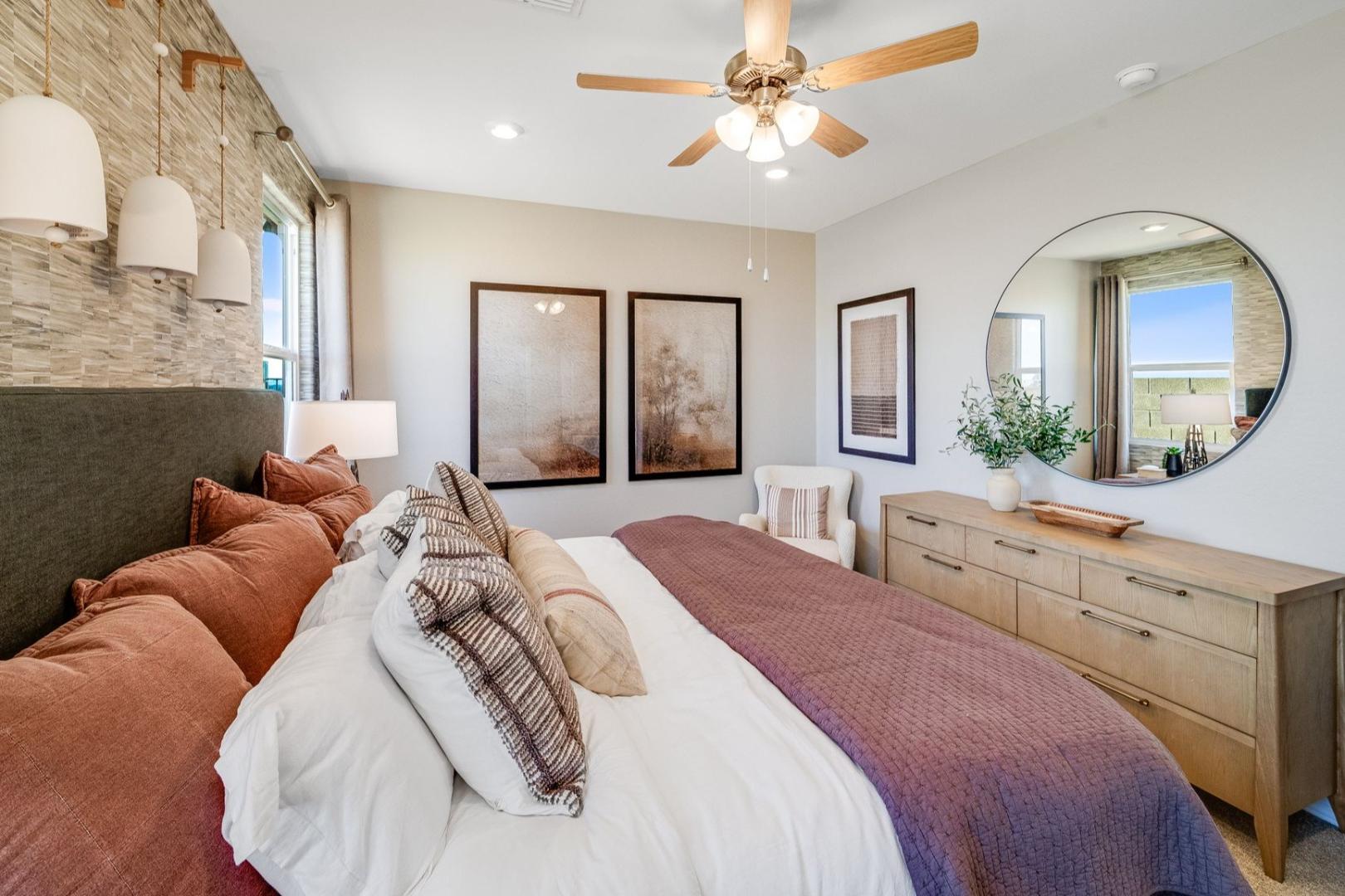Master suite in The Savannah B with plush king bed, neutral tones, ceiling fan, round mirror, and abstract art