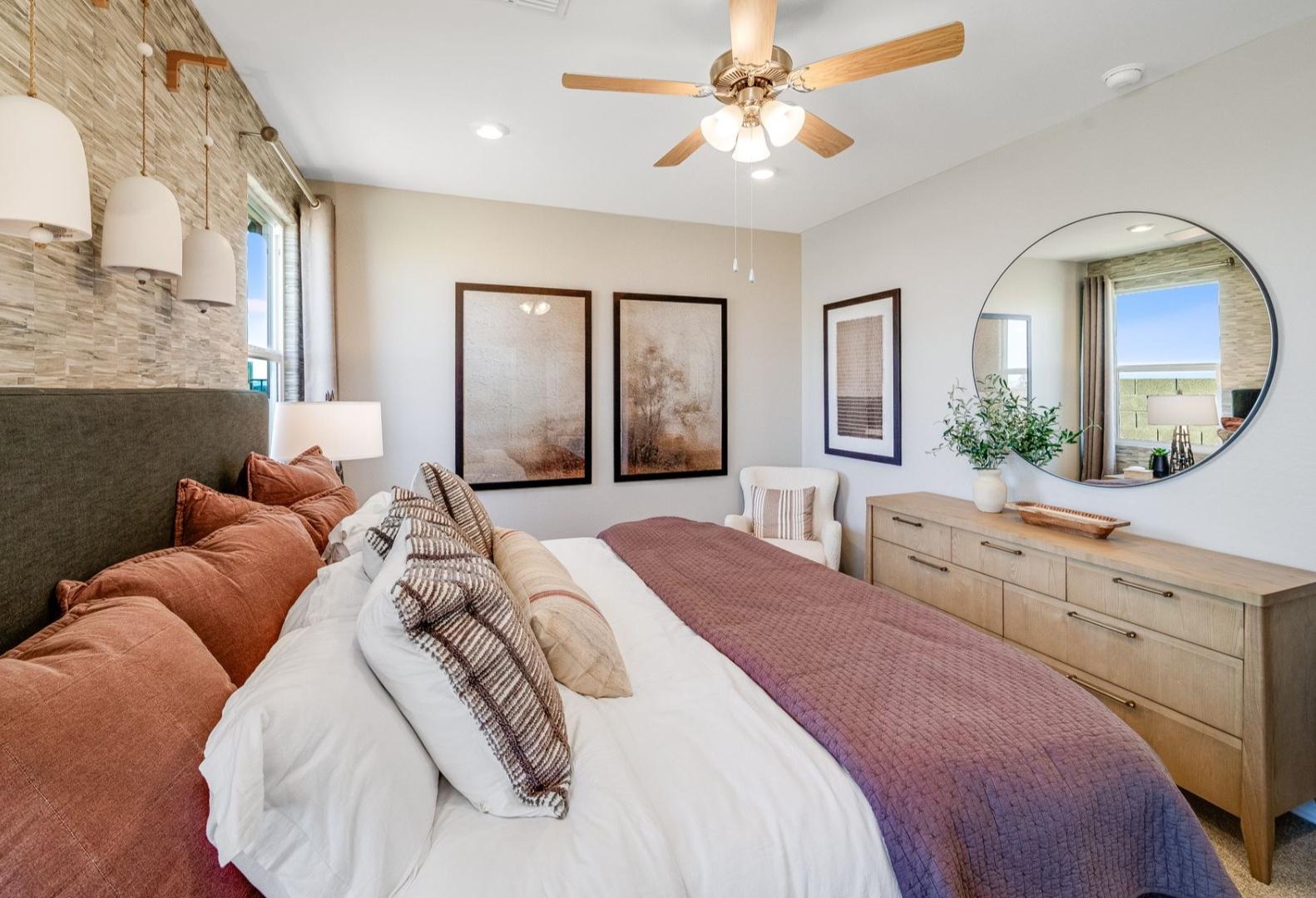Spacious master bedroom in The Savannah A with plush bed, wooden dresser, round mirror, and abstract art accents